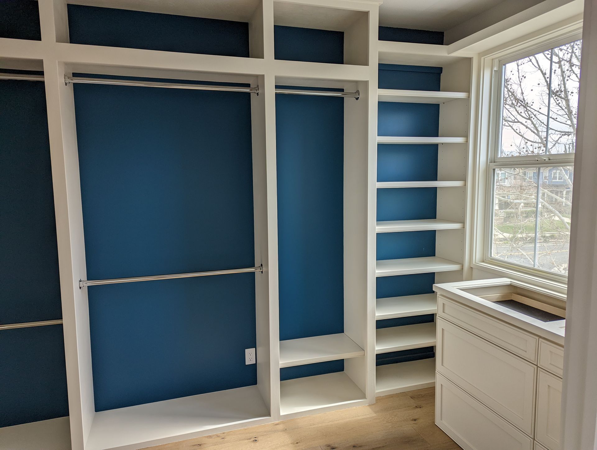 Built-in closet with blue walls, white shelves, and a window beside a white cabinet.