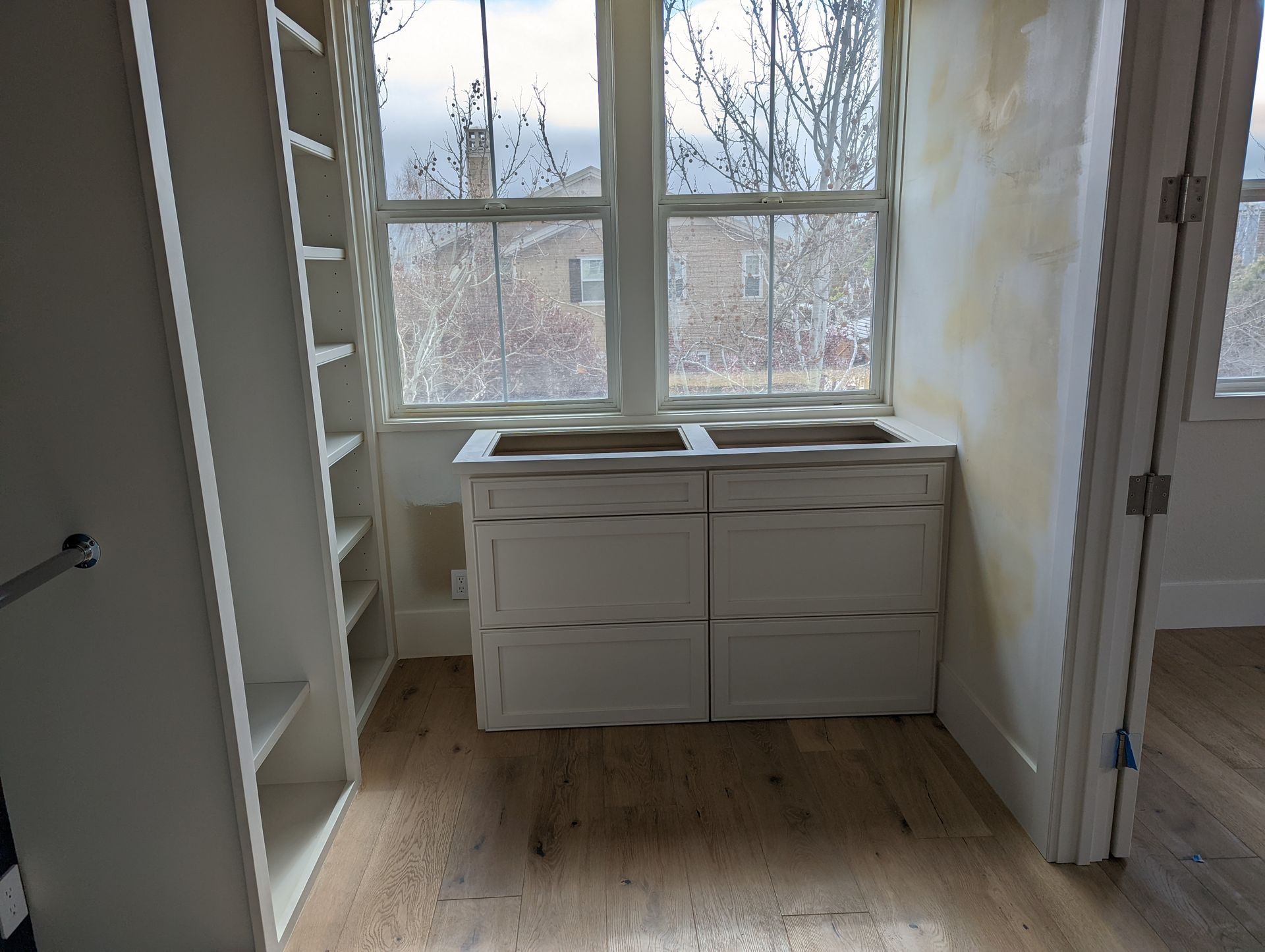 Empty room with white cabinets under a window, shelving on the left, and a door on the right.