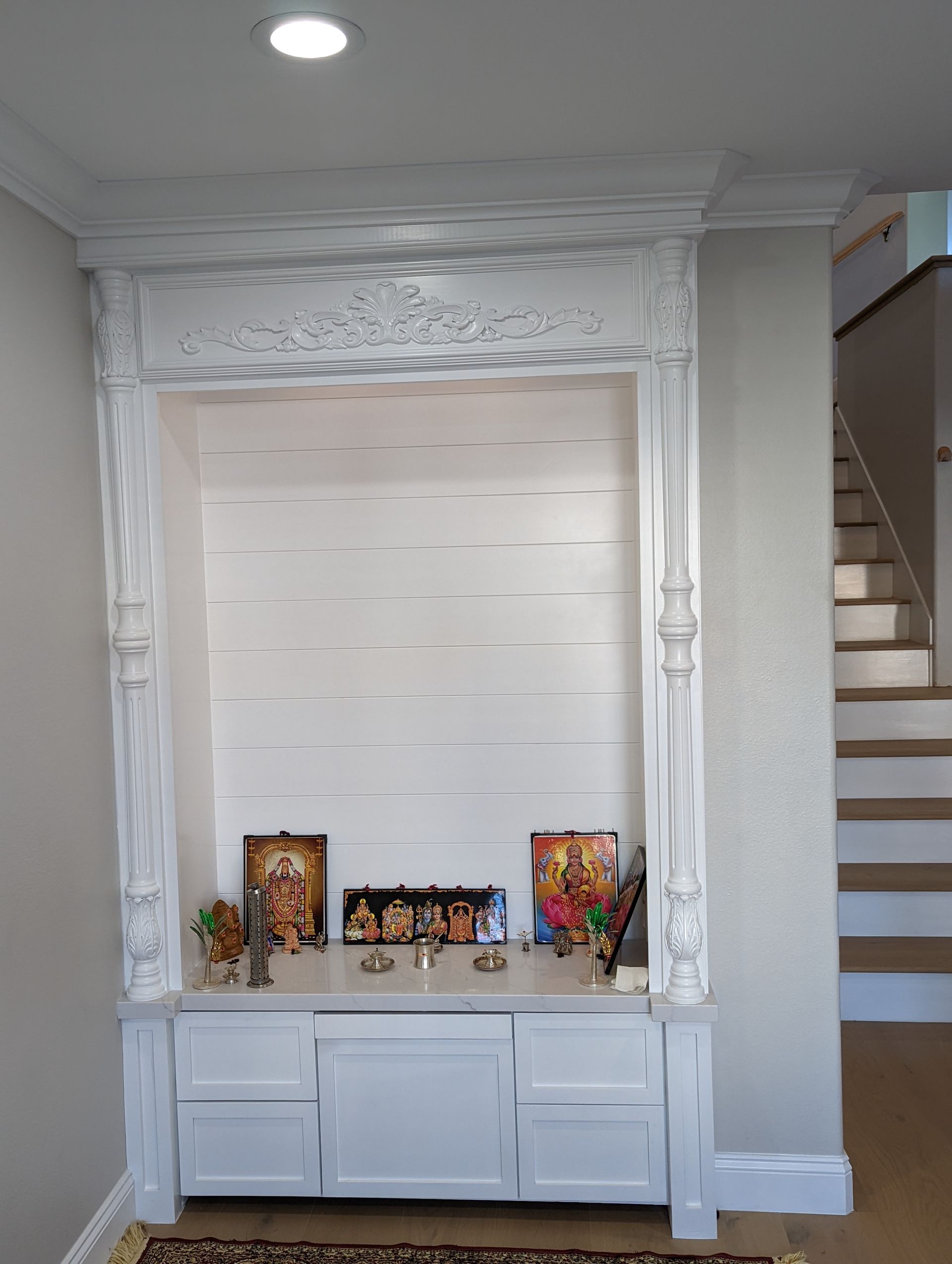 White home altar niche with framed icons, candles, and a cabinet beneath a decorative carved arch