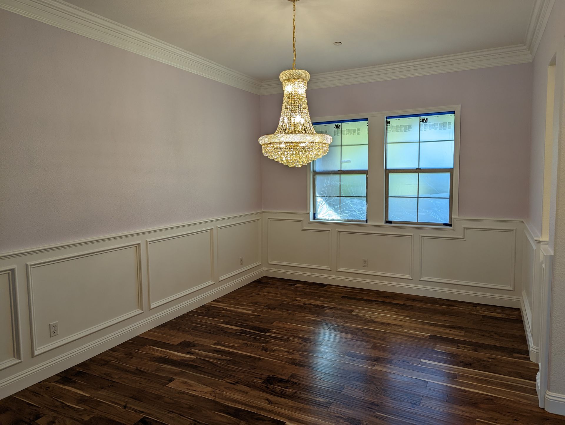 Empty dining room with chandelier, white paneled walls, and dark hardwood floors, lit by a window.