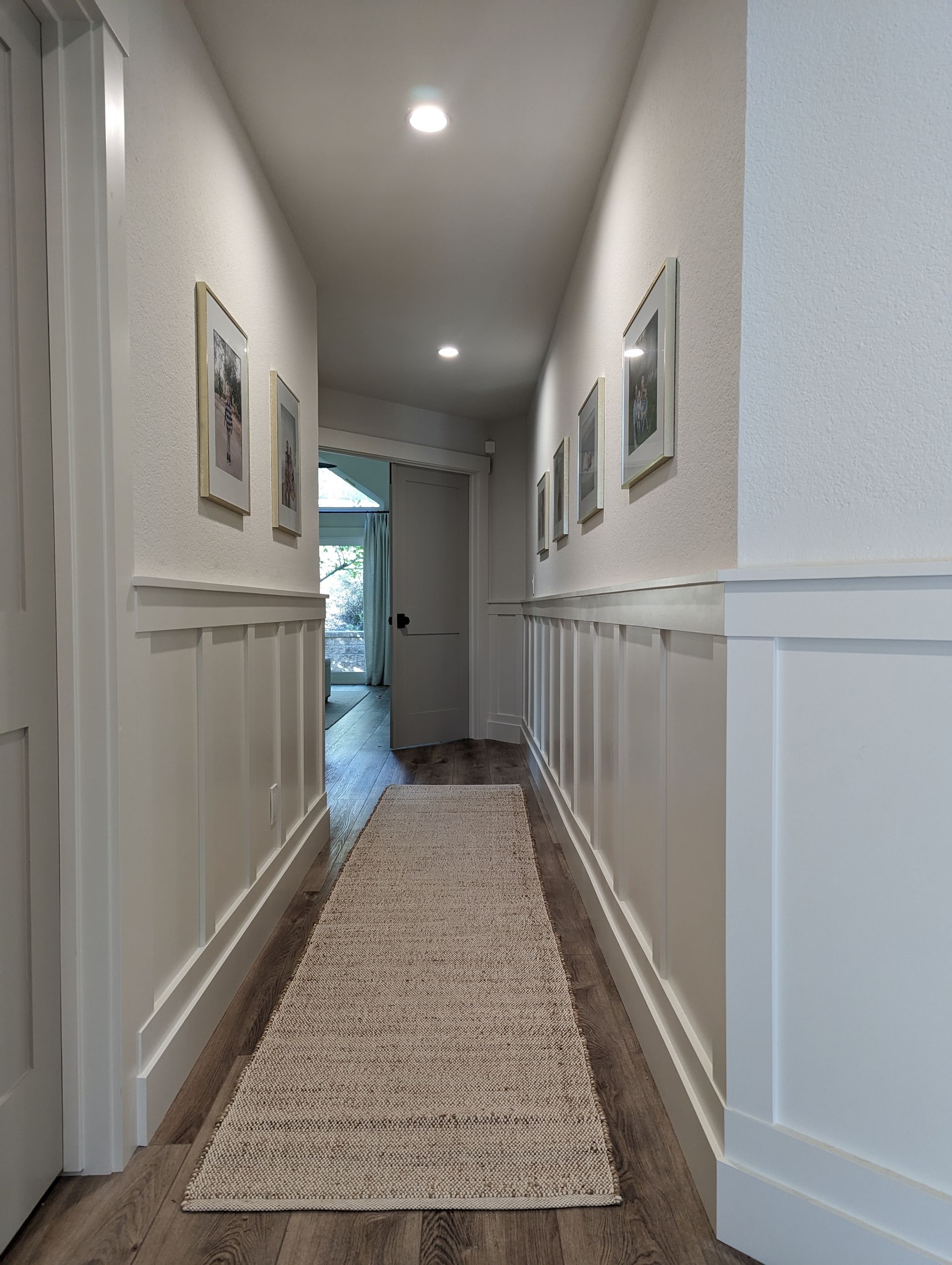 Narrow white hallway with framed art, recessed lights, and a long patterned runner rug leading to a doorway.