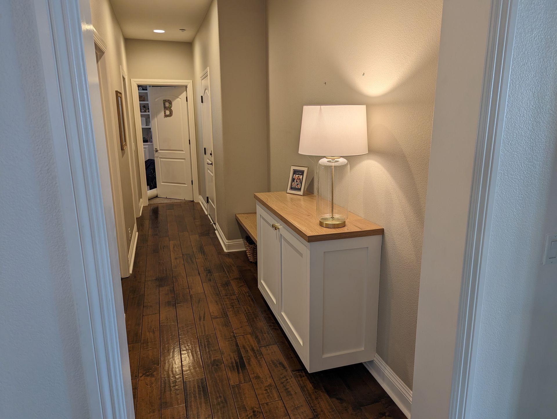 Narrow hallway with dark wood floor, white walls, and a lit lamp on a small cabinet.