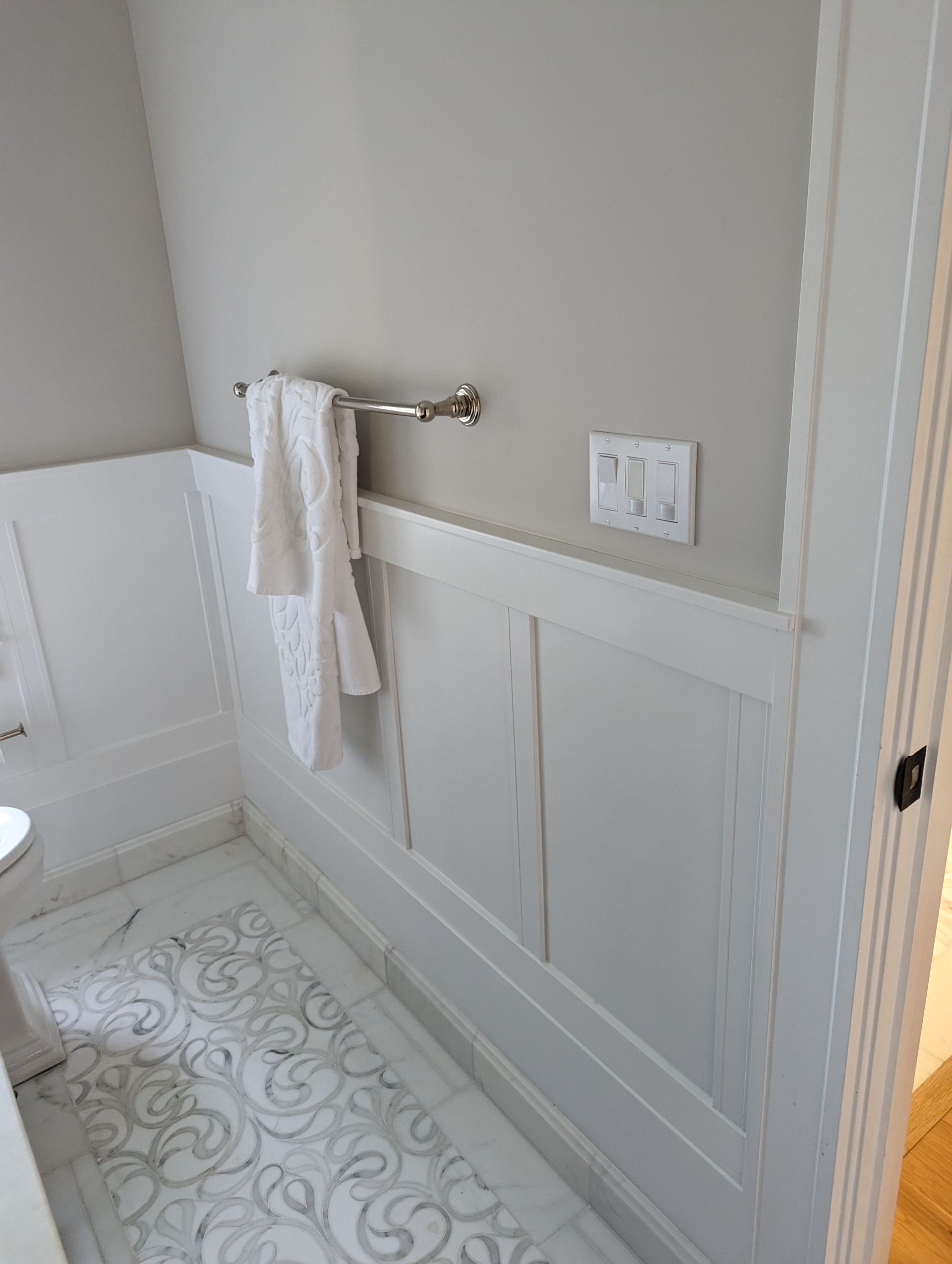 Small bathroom wall with towel rack, white wainscoting, outlet, and light patterned tile floor.