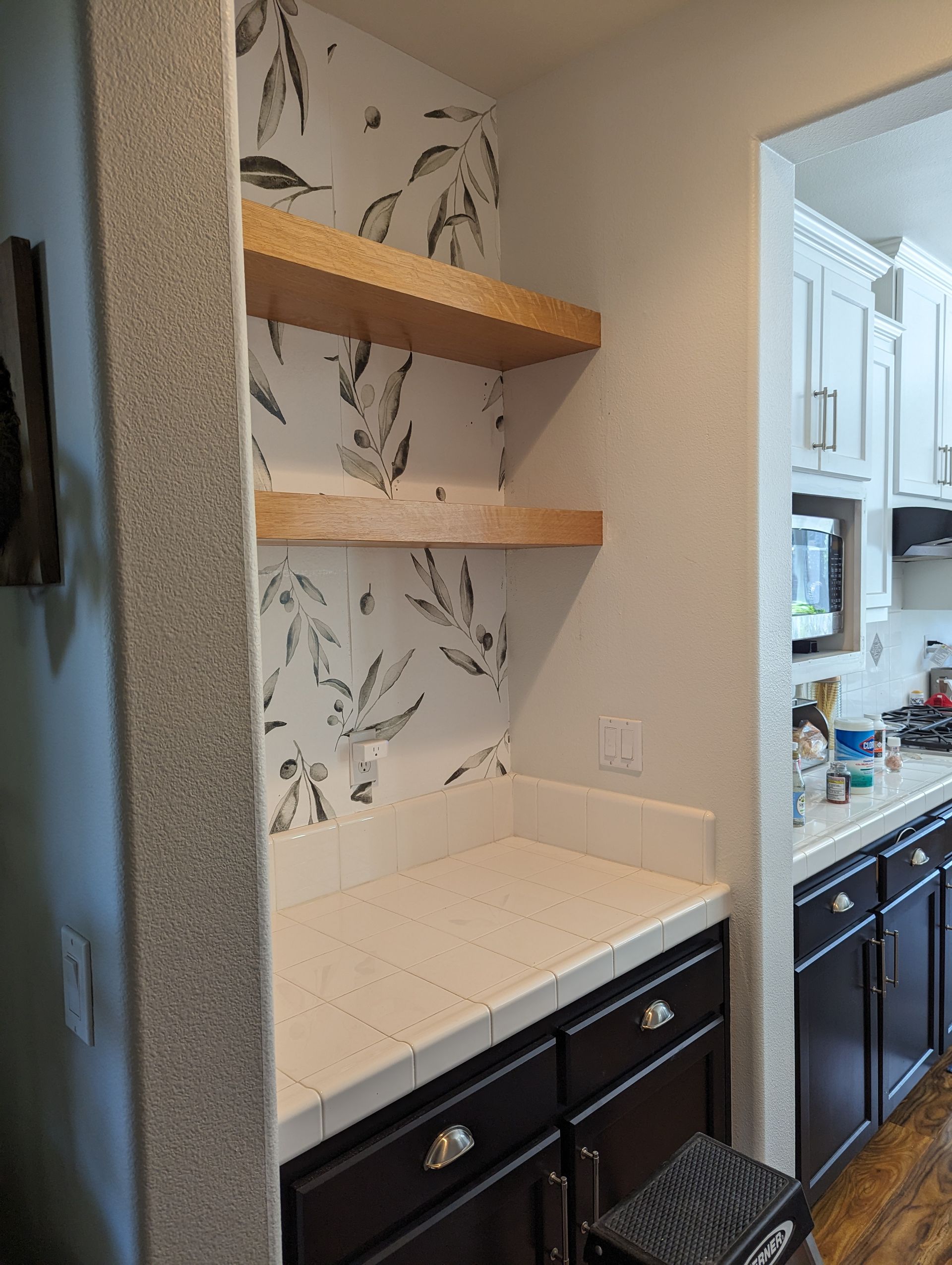 Kitchen nook with white countertop, dark cabinets, open wood shelves, and patterned wallpaper