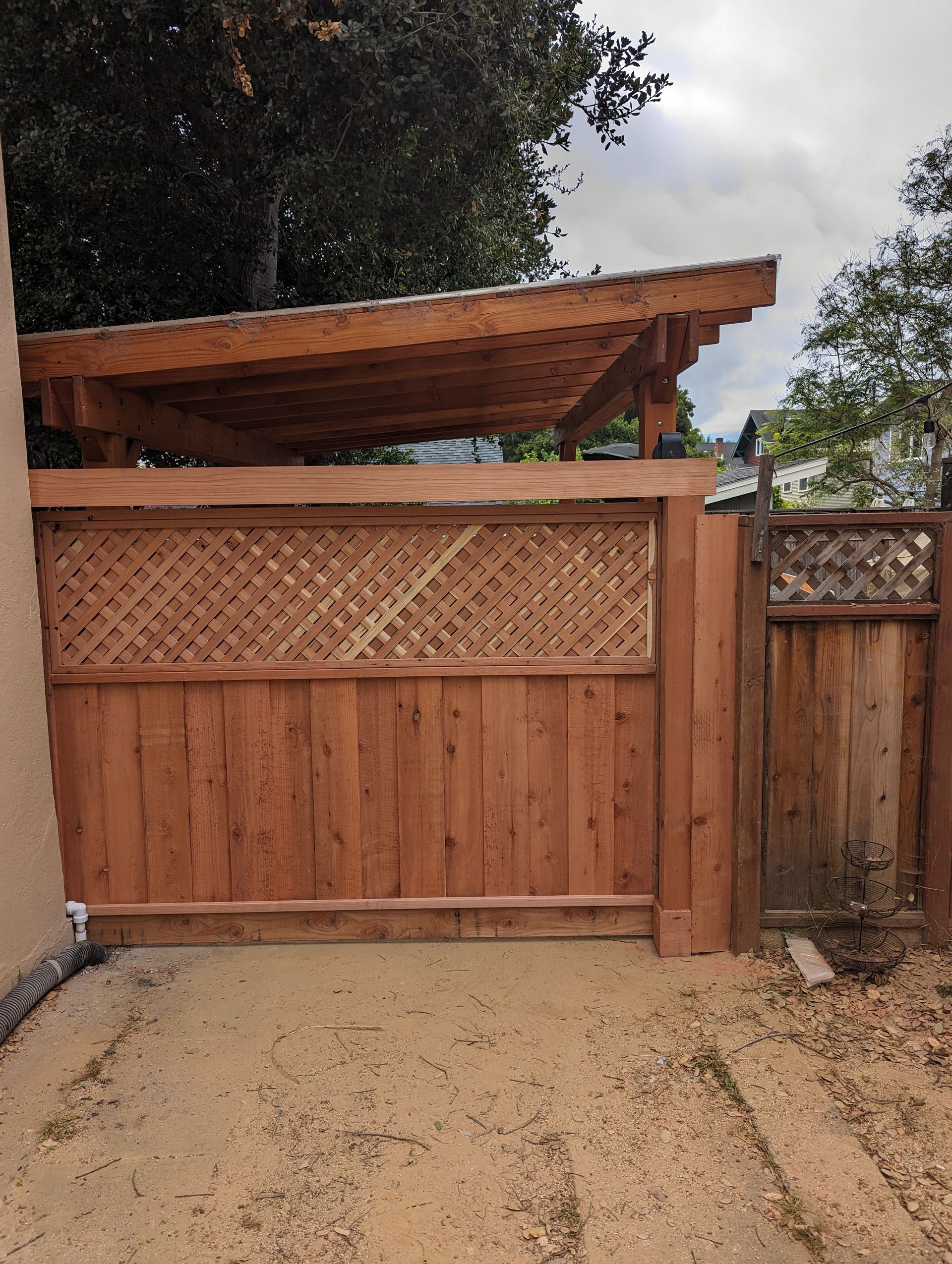 Wooden driveway gate with lattice top under a slanted roof, beside a fence and dirt path.