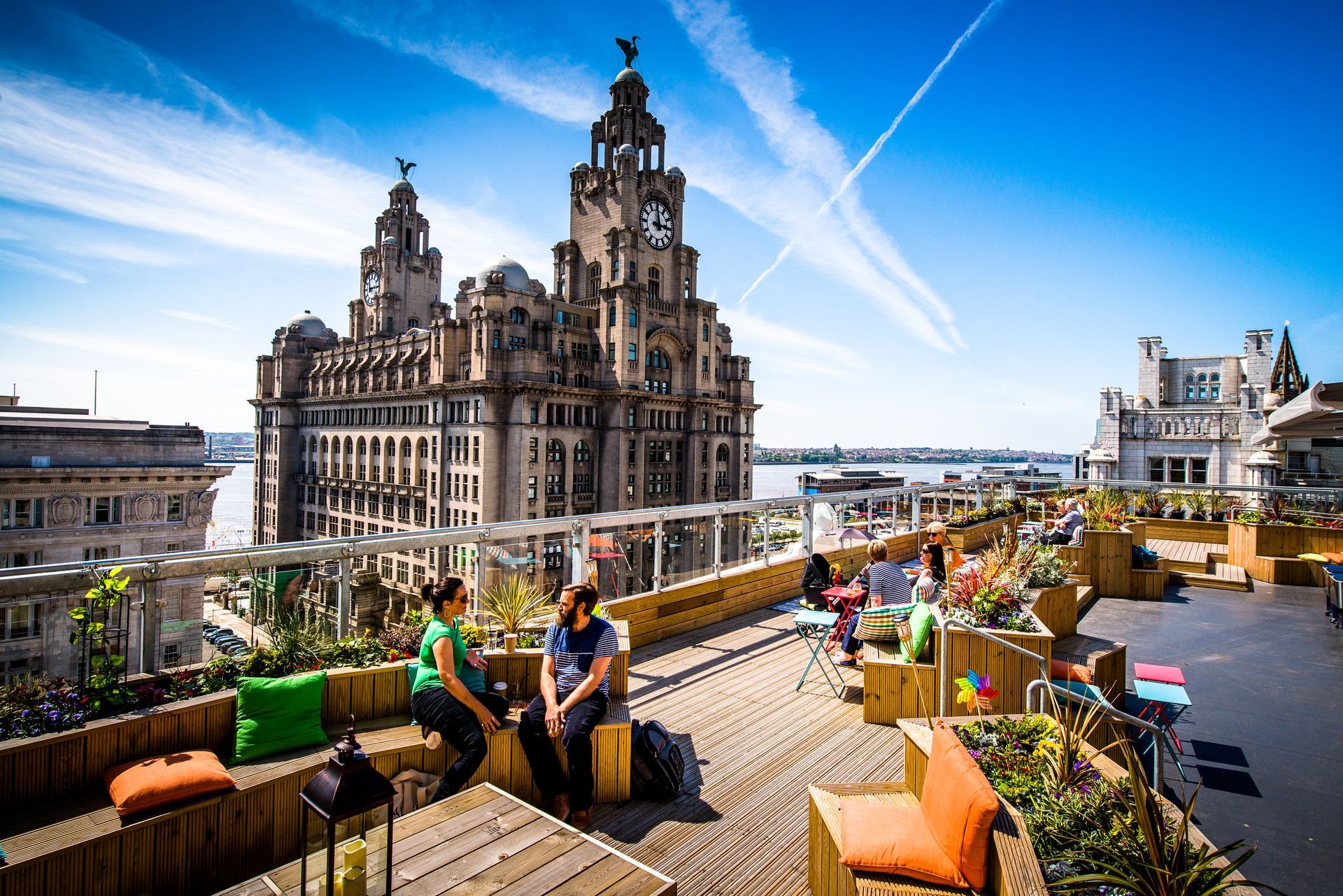 Rooftop bar with people, wooden seating, lush plants, overlooking Liver Building in Liverpool, UK. Sunny day.