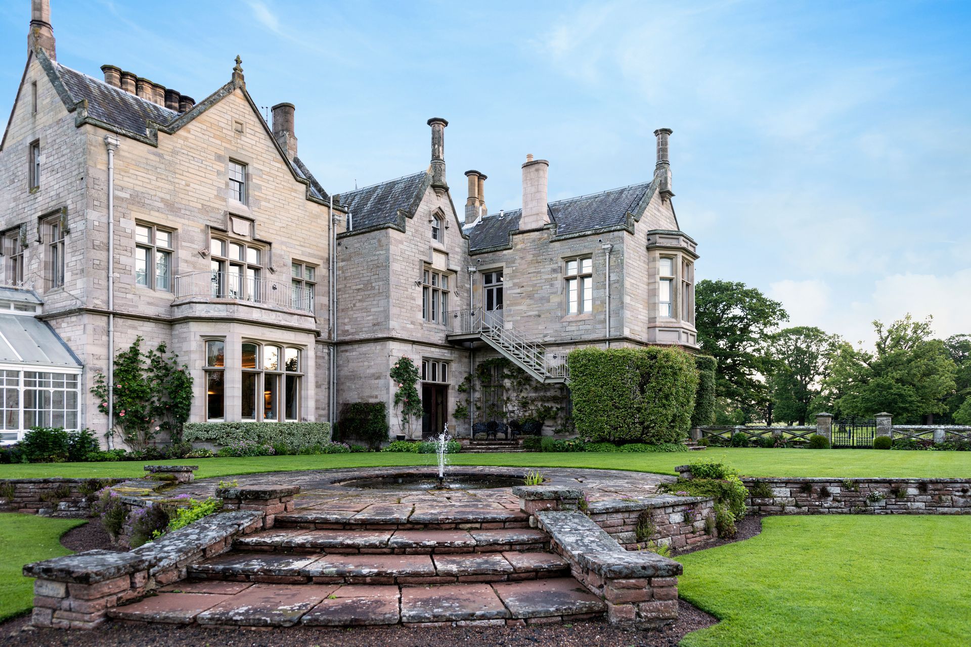 Stone manor house with a tiered garden, green lawn, and fountain. Blue sky in background.