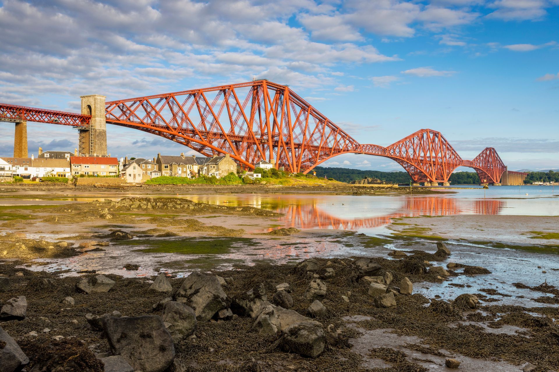 Red Forth Bridge over water; houses in background under blue sky.