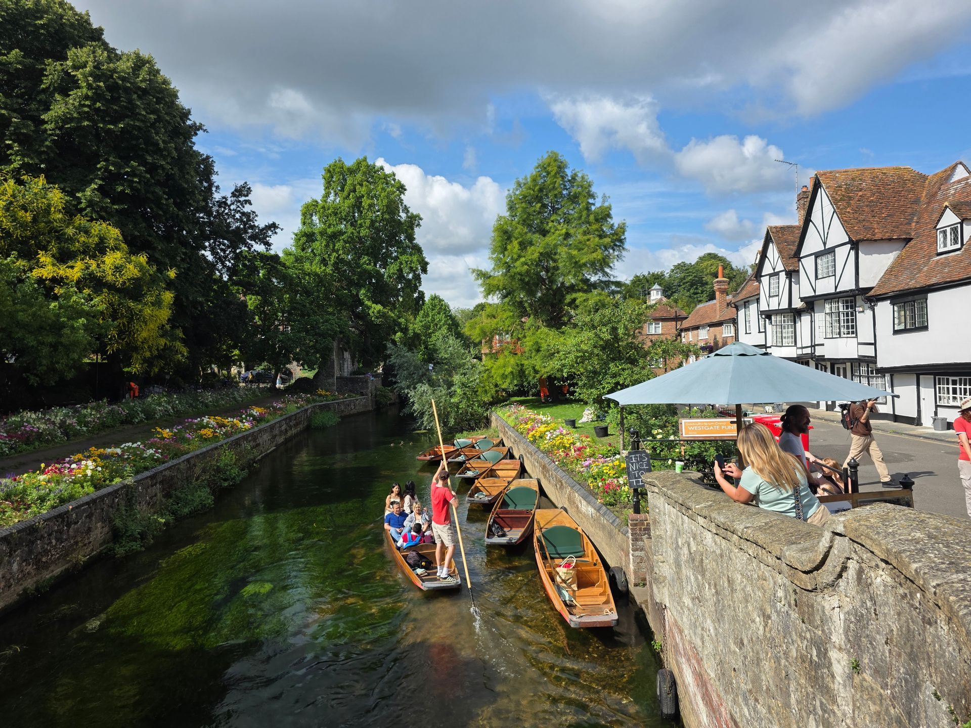 Punts on a river lined with flowers. Historic buildings and people under an umbrella are visible. Sunny day.