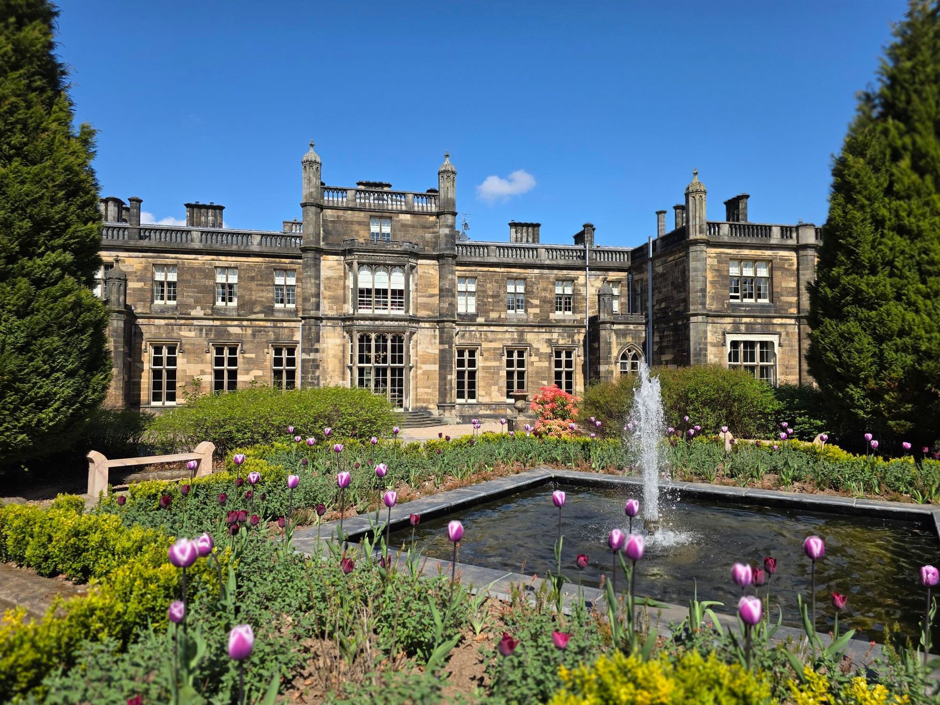 Stone mansion with a fountain and flower garden under a blue sky.