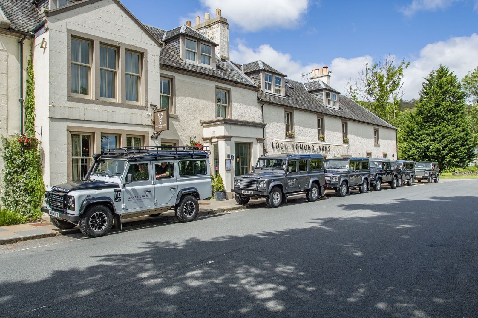 Row of gray Land Rovers parked outside a light-colored inn under a blue sky.