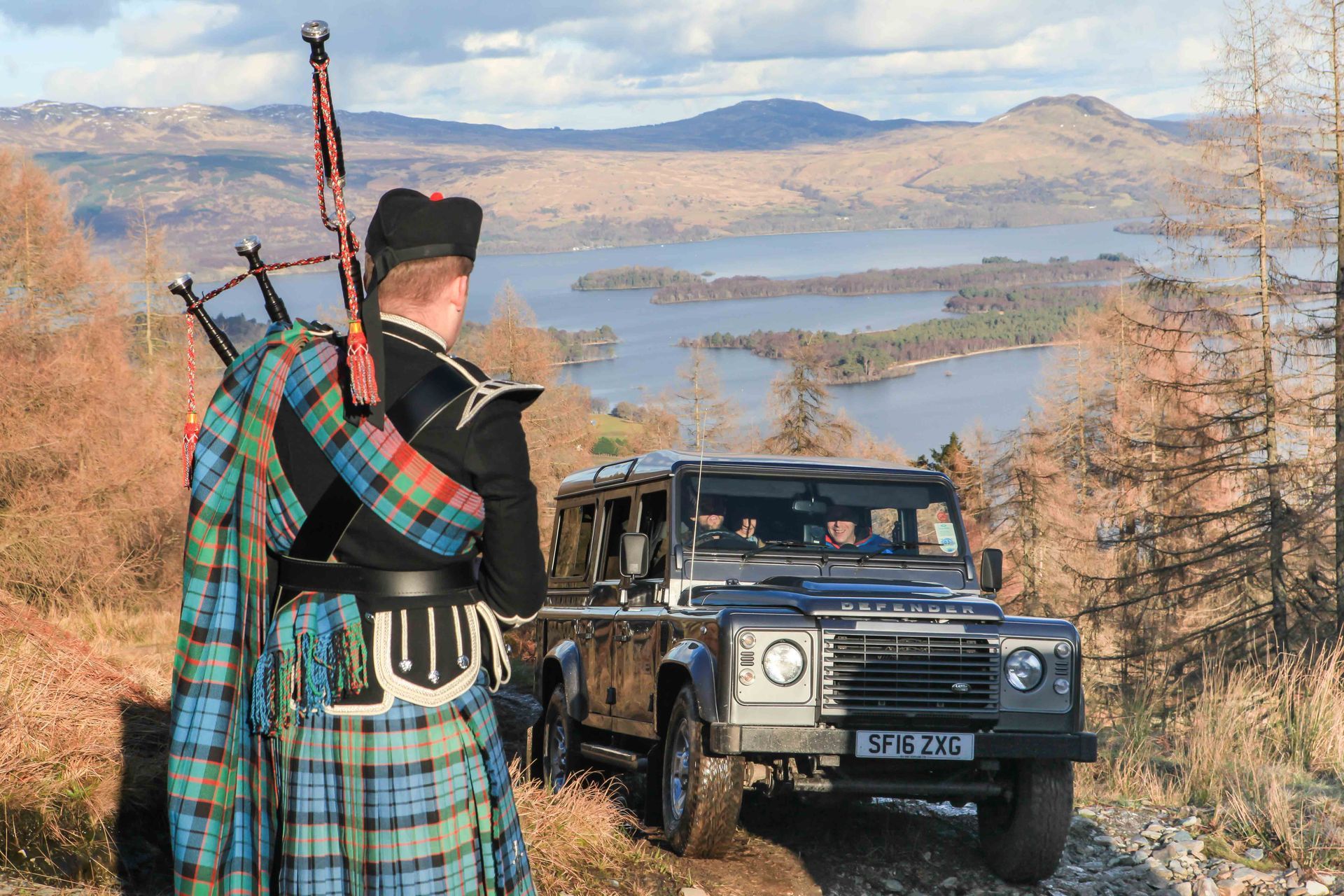 Bagpiper in traditional attire stands near a Land Rover with a scenic loch and mountains in the background.