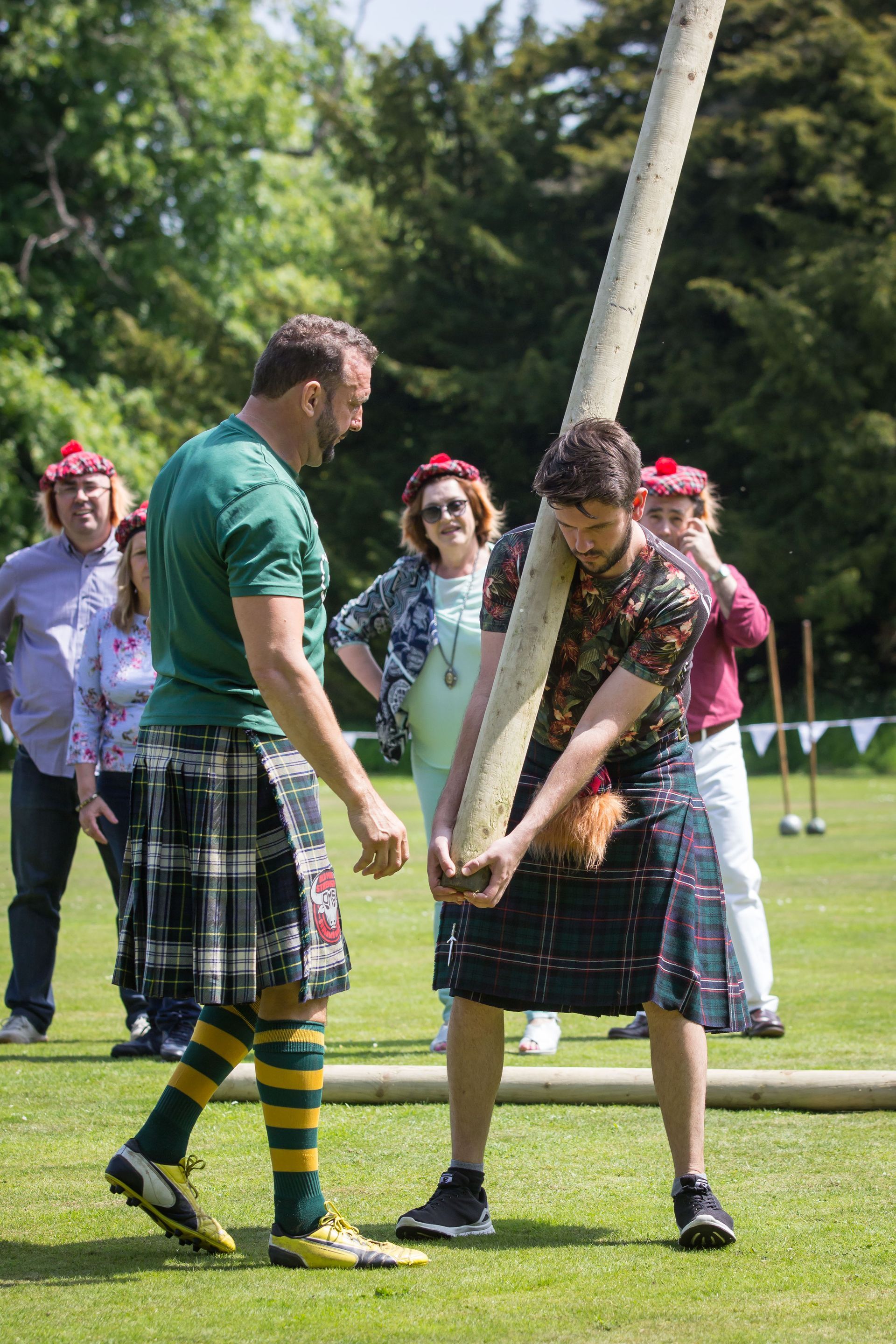 Two men in kilts hefting a large log in an outdoor competition. Spectators watch on a grassy field.