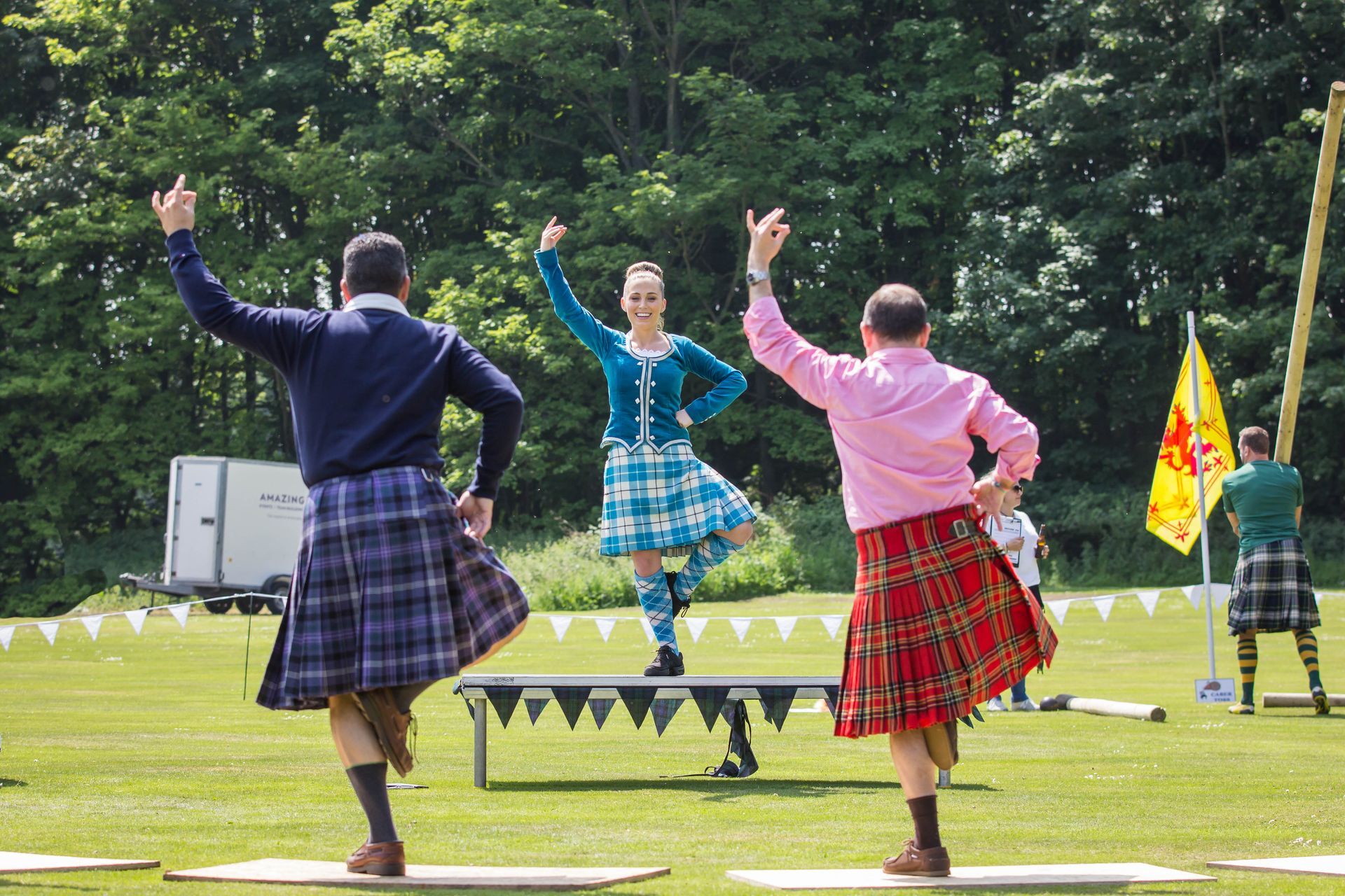 Scottish dancers in kilts performing on grass, arms raised. A yellow flag is held by someone in the background.