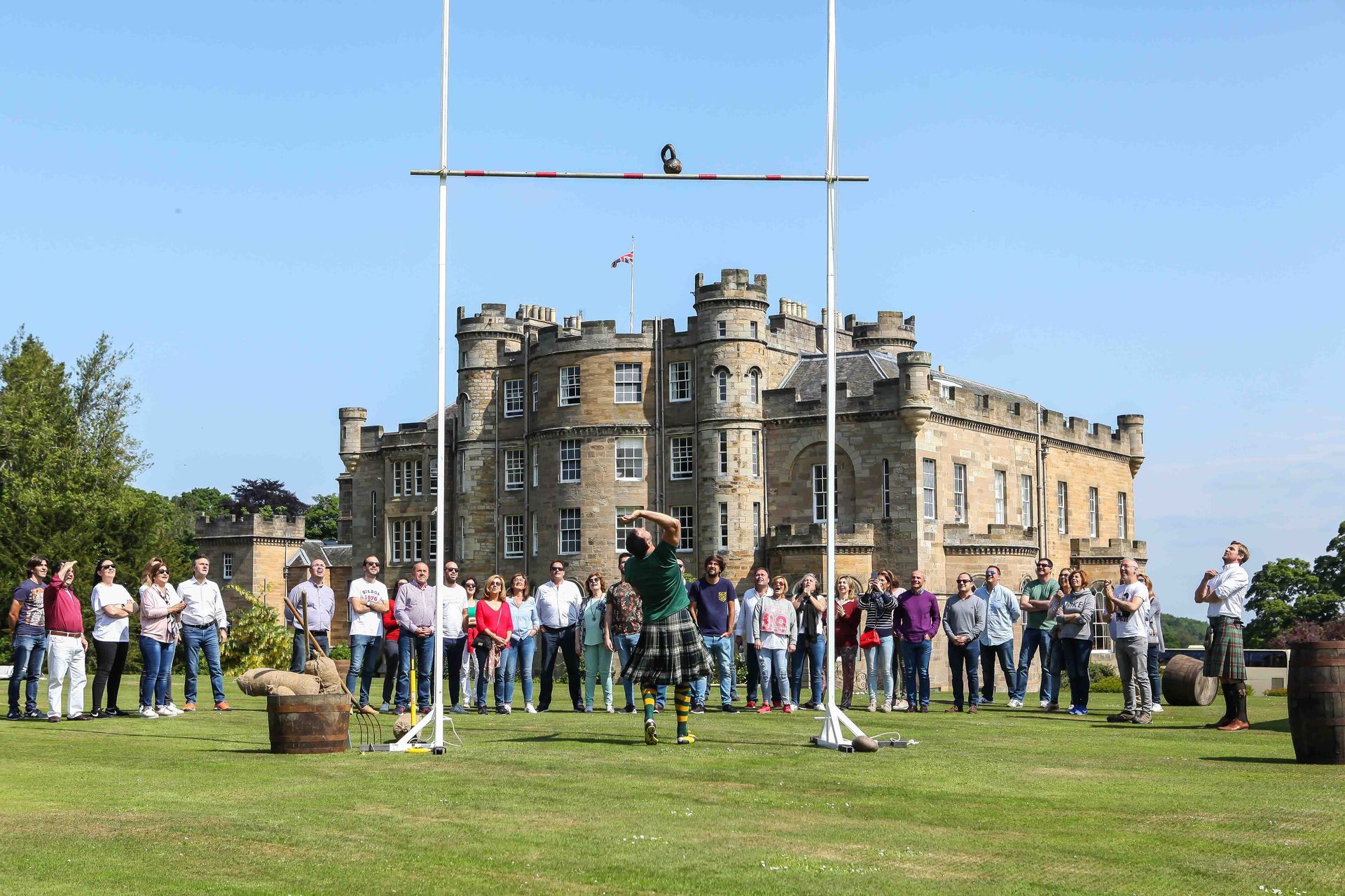 A man in a kilt tosses a caber at a Highland Games event in front of a castle. Spectators watch.