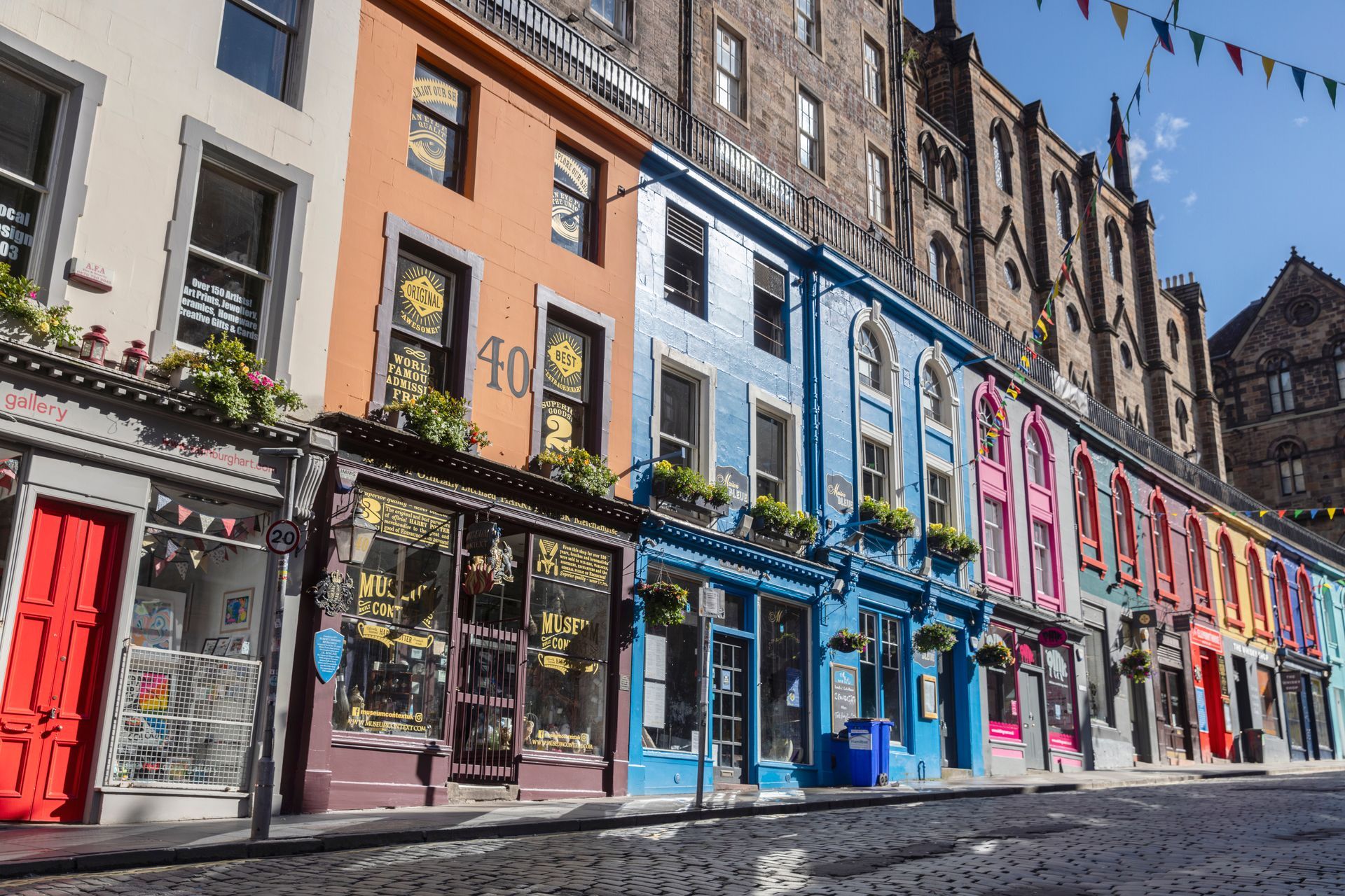 Colorful shops along a cobblestone street in Edinburgh, Scotland; buildings painted red, orange, blue, and pink.