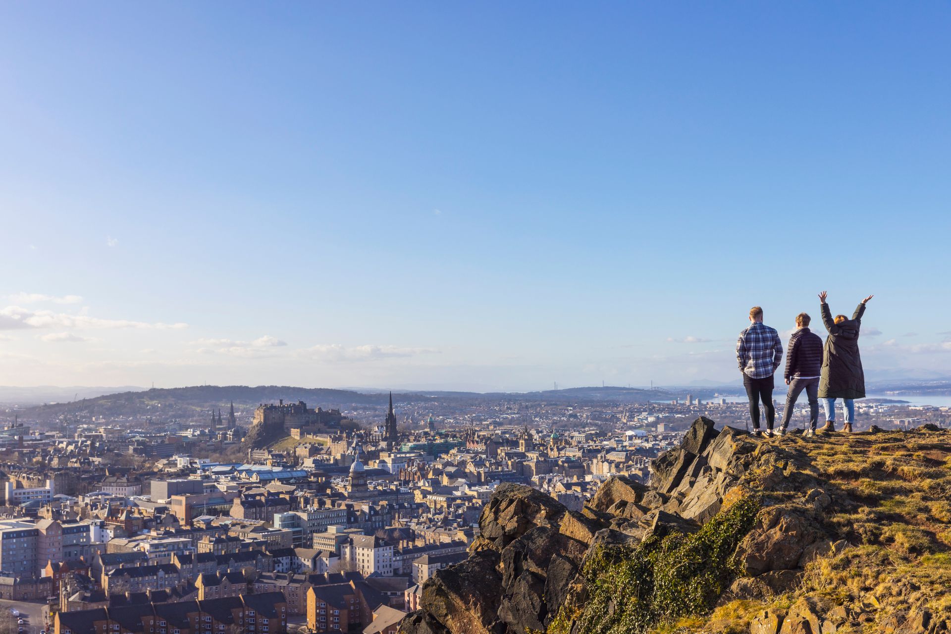 Three people overlooking a cityscape, one with arms raised, on a sunny day.