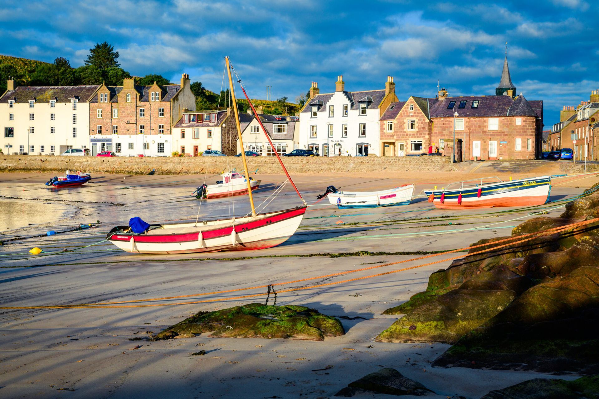 Boats sit on a sandy beach in front of a row of buildings. Blue sky overhead.