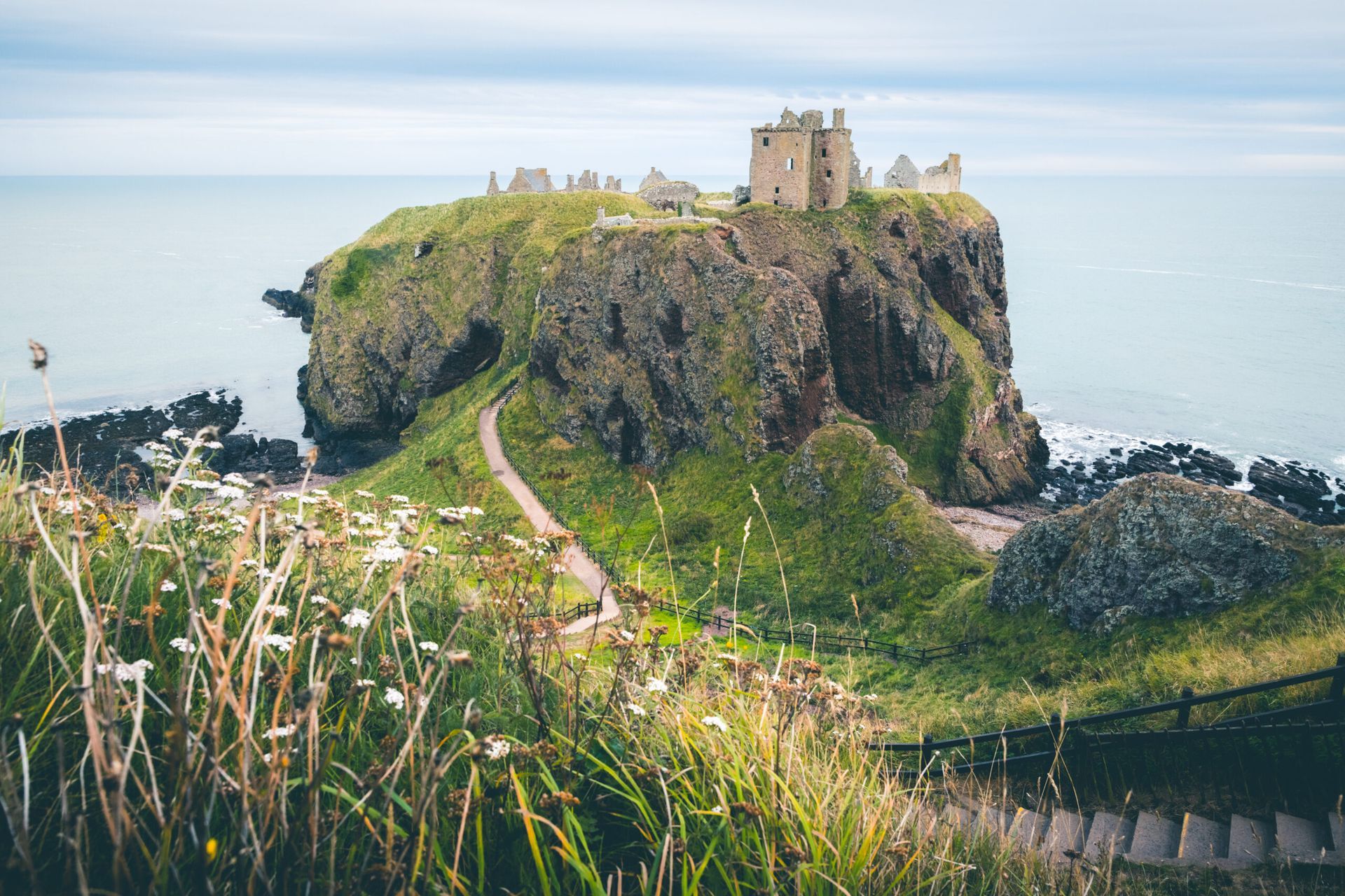 Stone castle ruins atop a rocky cliff overlooking the ocean, with a path leading to it.