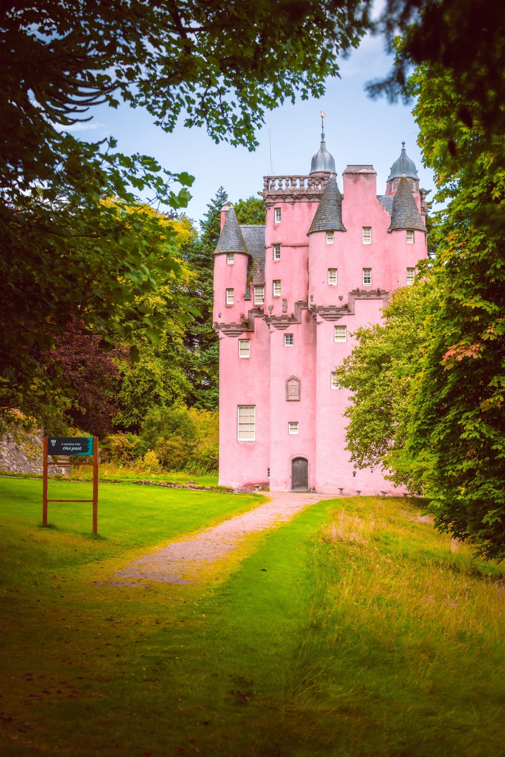 Pink castle surrounded by green trees, pathway leading to the entrance, blue sky.