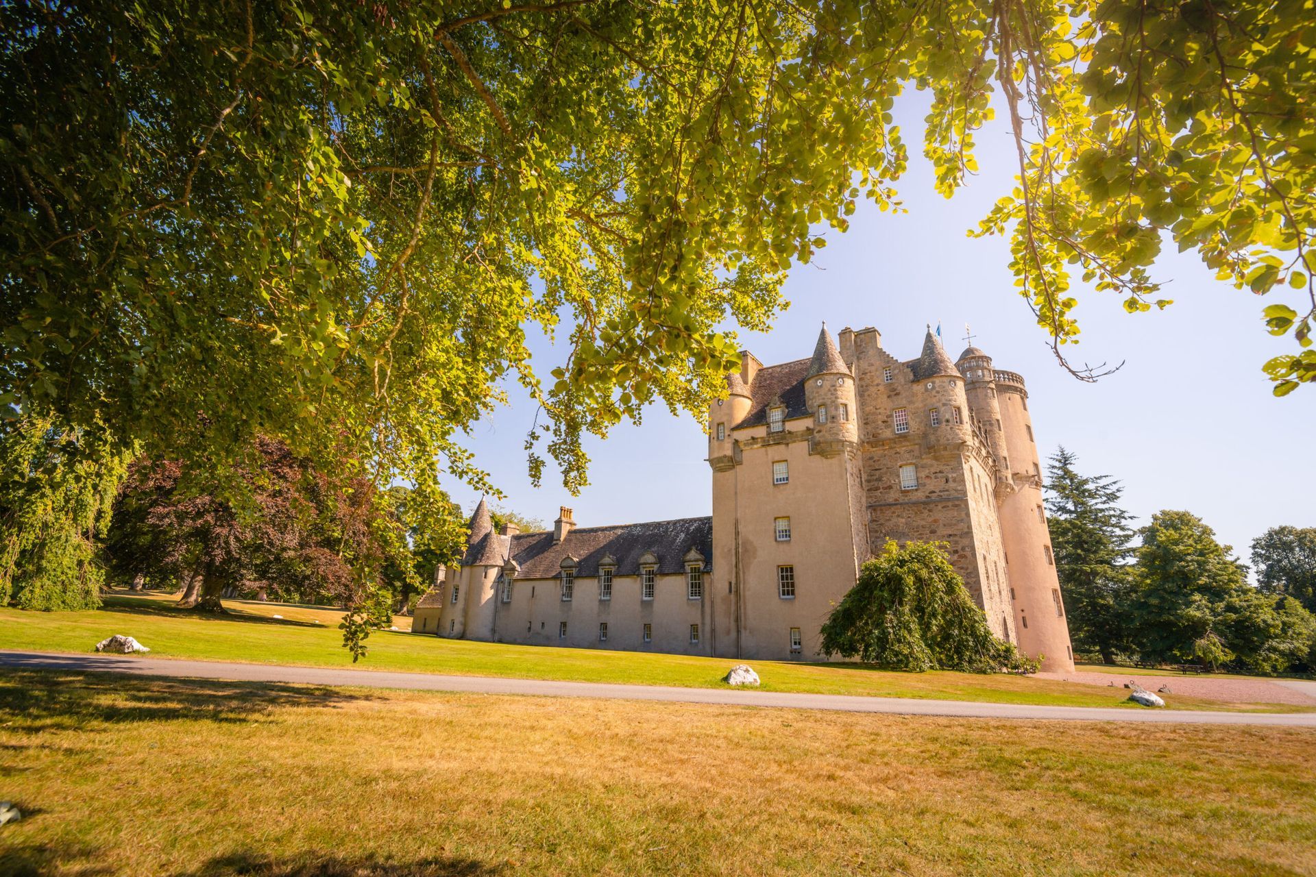 Scottish castle with tower, on grassy lawn under trees. Sunny day.