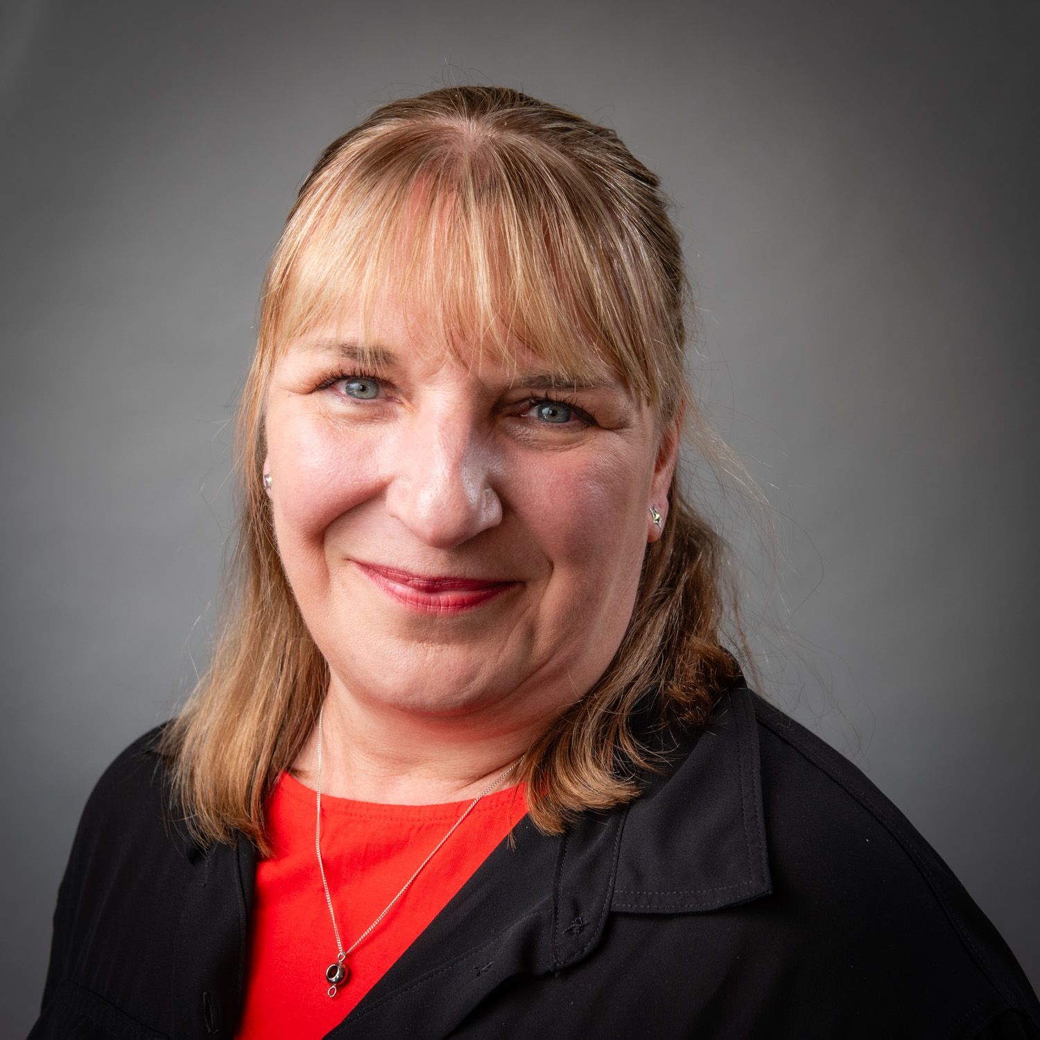 Woman with blonde hair, smiling at the camera, wearing a red top and black jacket. Gray background.