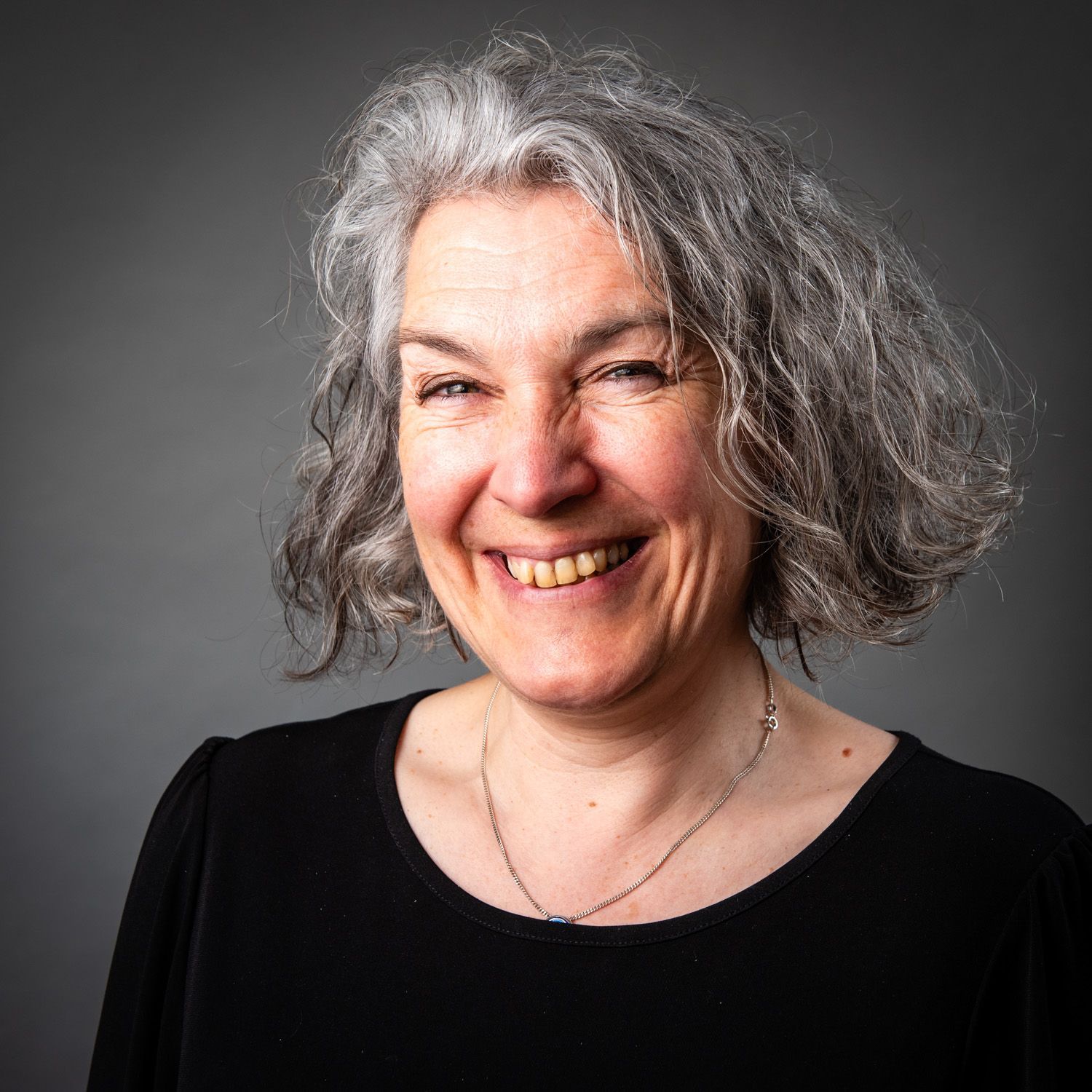 Woman with short, gray curly hair, smiling widely, wearing a black top. Gray background.