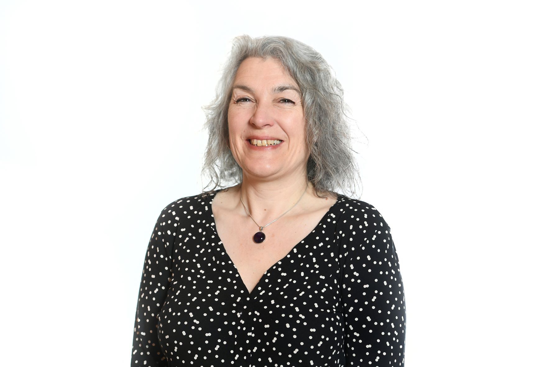 Woman with gray hair smiling, wearing a black and white polka dot top, against a white background.
