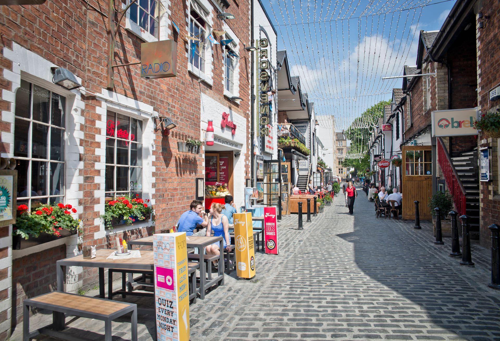 Cobblestone street lined with brick buildings, outdoor seating at restaurants, and pedestrians.