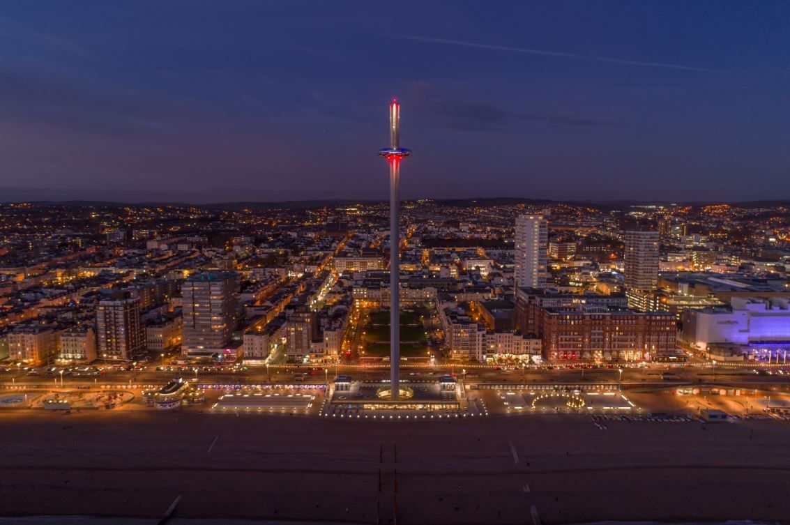 Tall, illuminated tower over Brighton, England at dusk. City lights and beach in foreground.