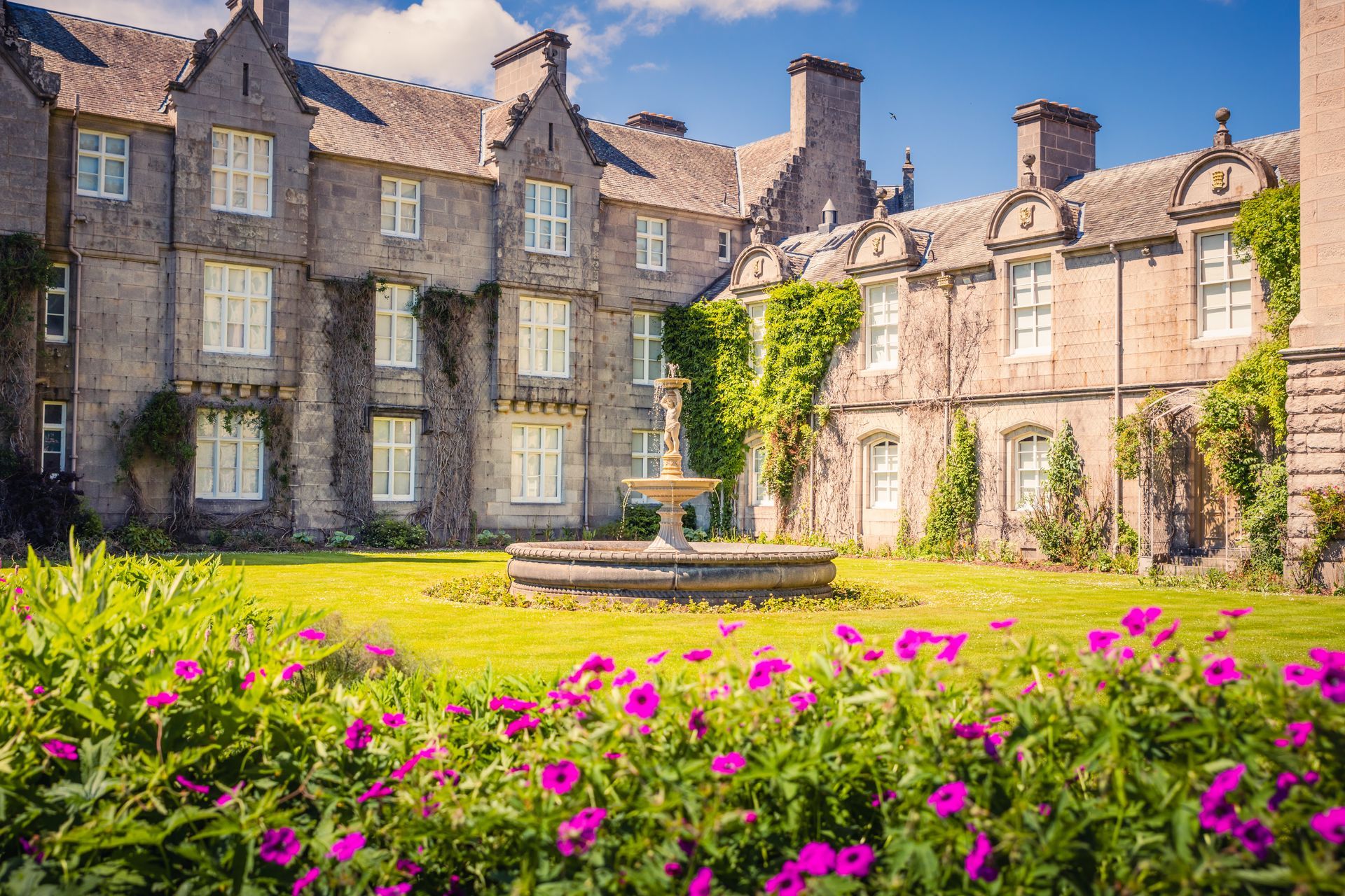 Exterior of a stone manor house with a fountain, lush lawn, and flowerbeds. Bright, sunny day.