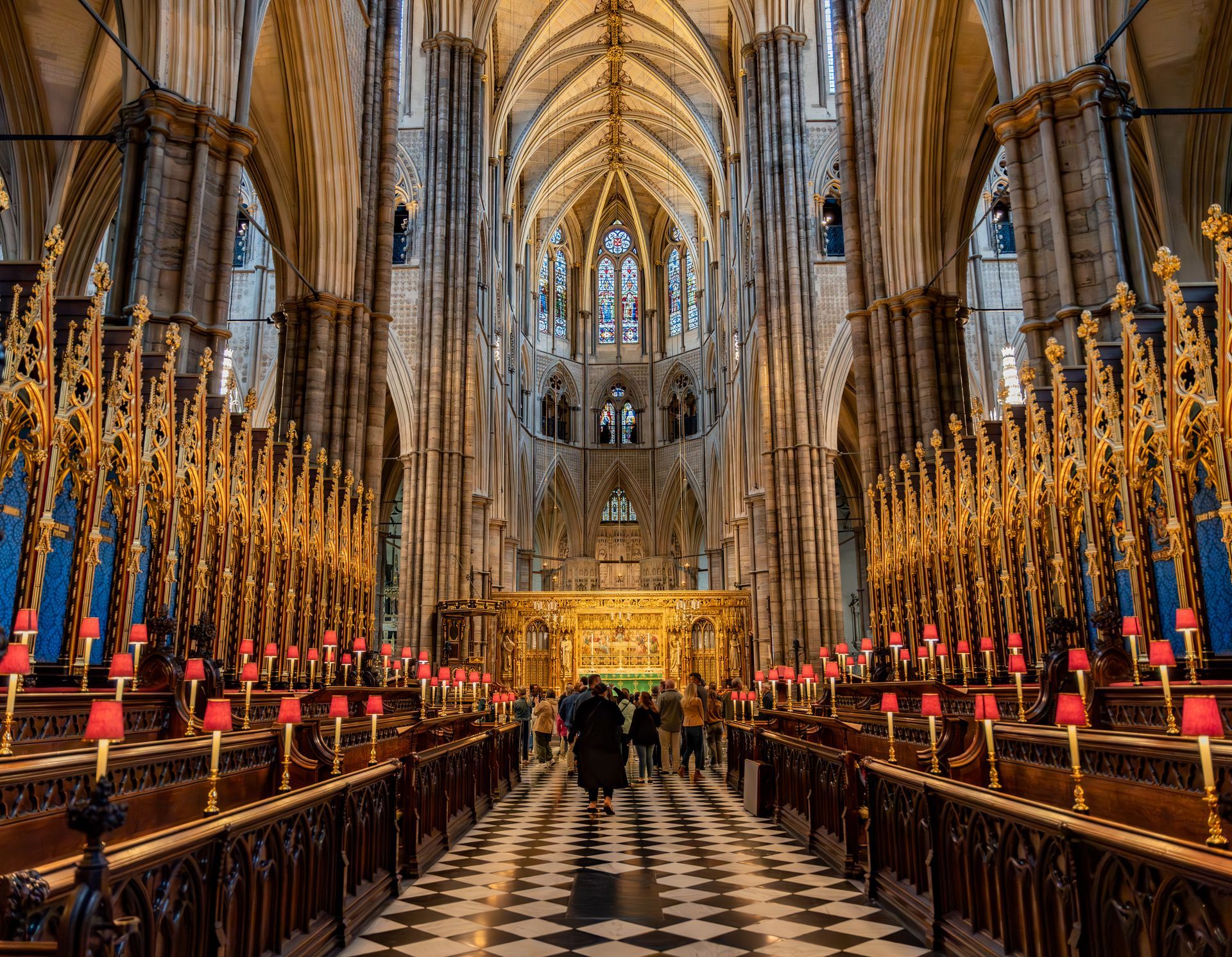 Interior of a cathedral, with arched ceilings and rows of ornate wooden pews. Several people walk towards the altar.