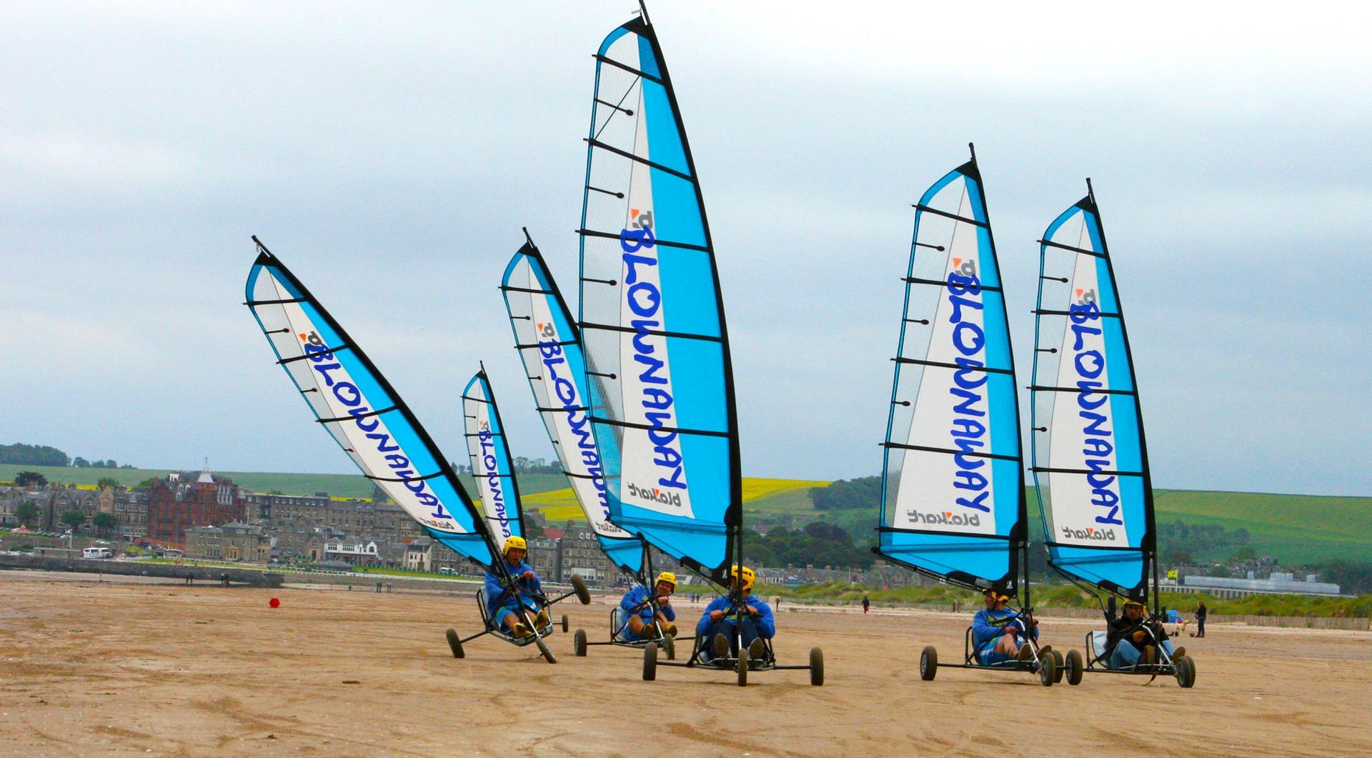Land yachts with blue sails race on a sandy beach.