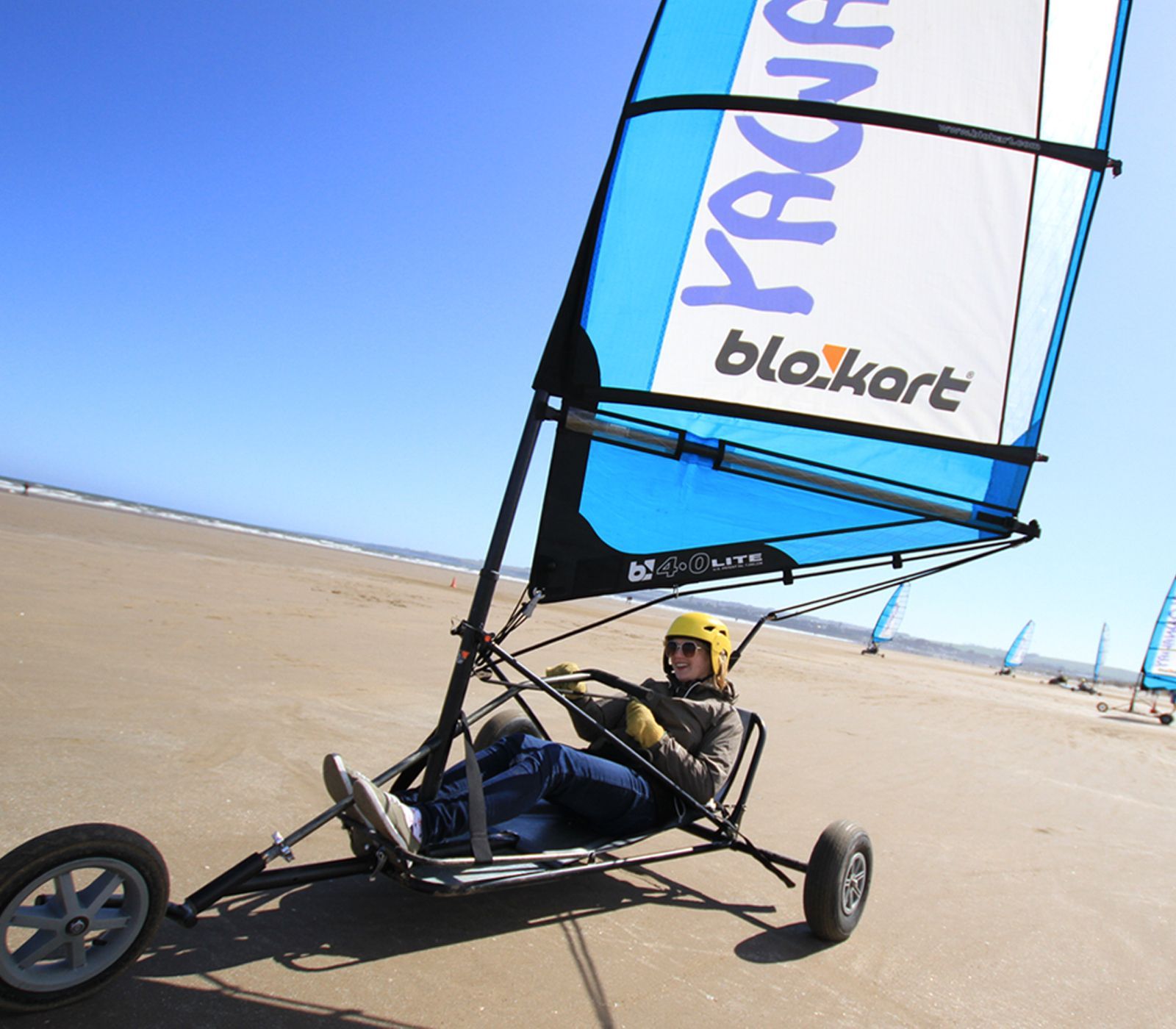 Person driving a land yacht with a blue and white sail on a sandy beach under a clear sky.