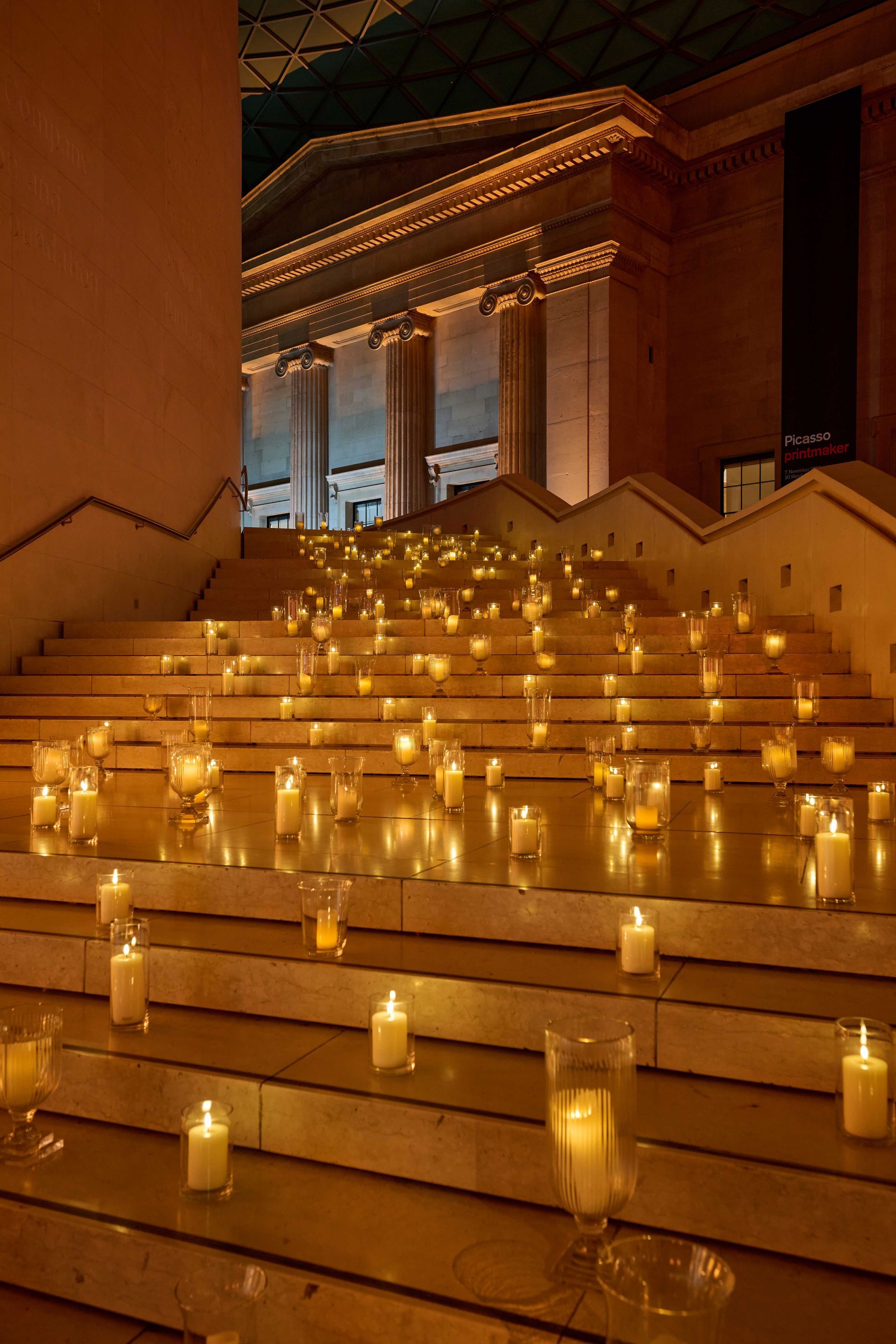 Candles illuminate a grand staircase in a building with columns; warm glow against a dark background.