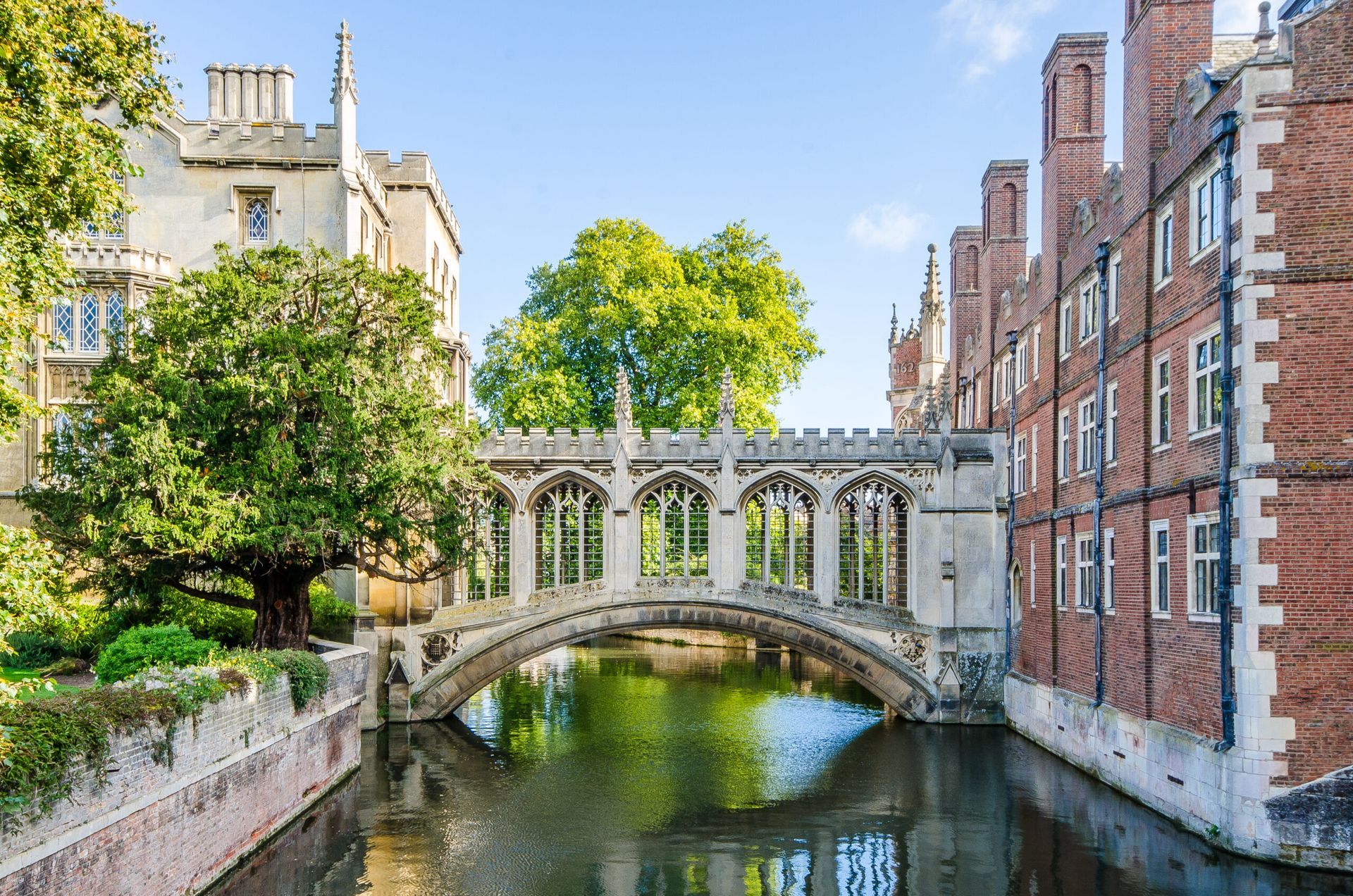 Cambridge's Bridge of Sighs over the River Cam, flanked by university buildings and greenery, under a blue sky.