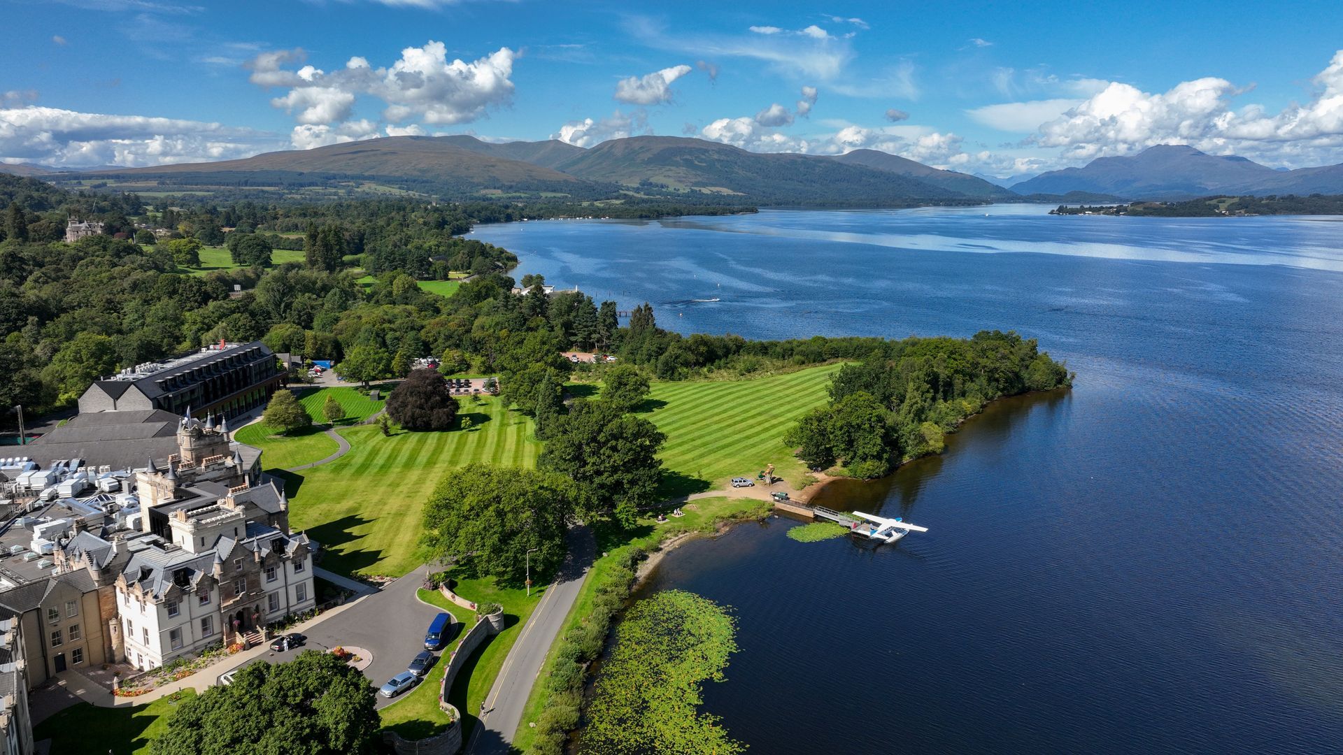 Aerial view of a lake and green landscape with a large building on the left. Blue sky and mountains in the background.