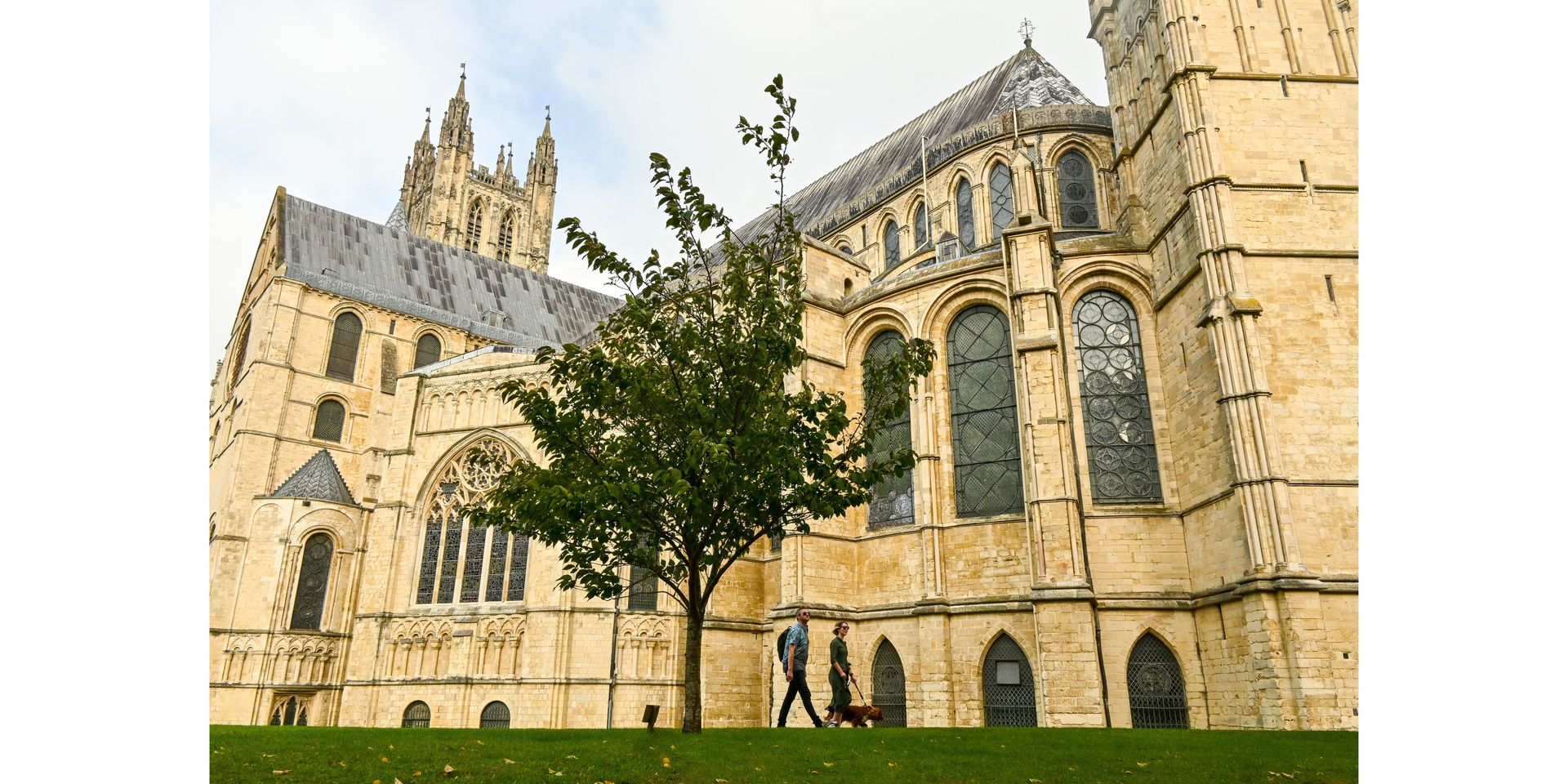 Canterbury Cathedral, a large beige stone building, with a tree in front. Two people walk on the grass.