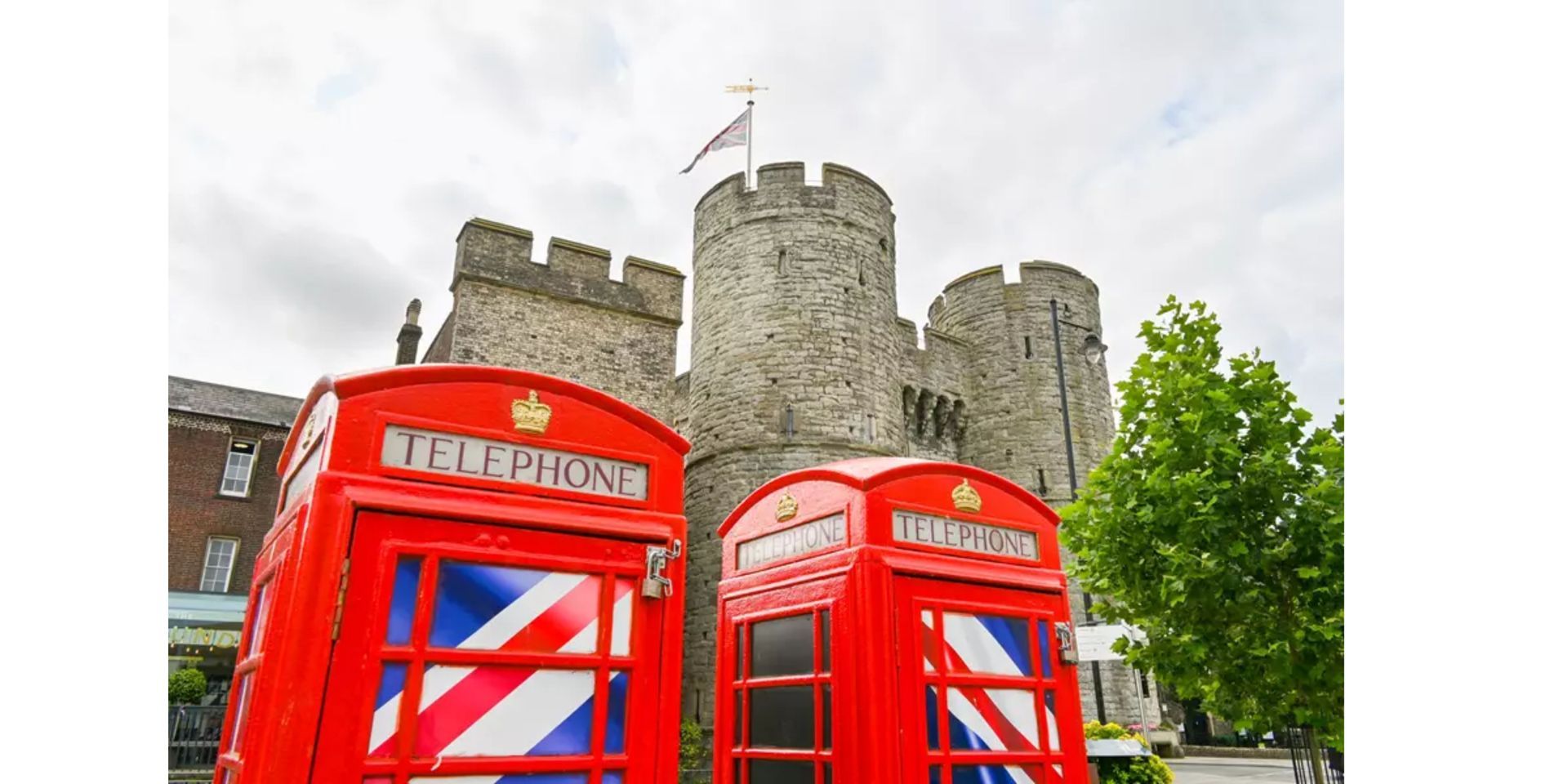 Two red British phone booths in front of a stone castle, with a flag on top.