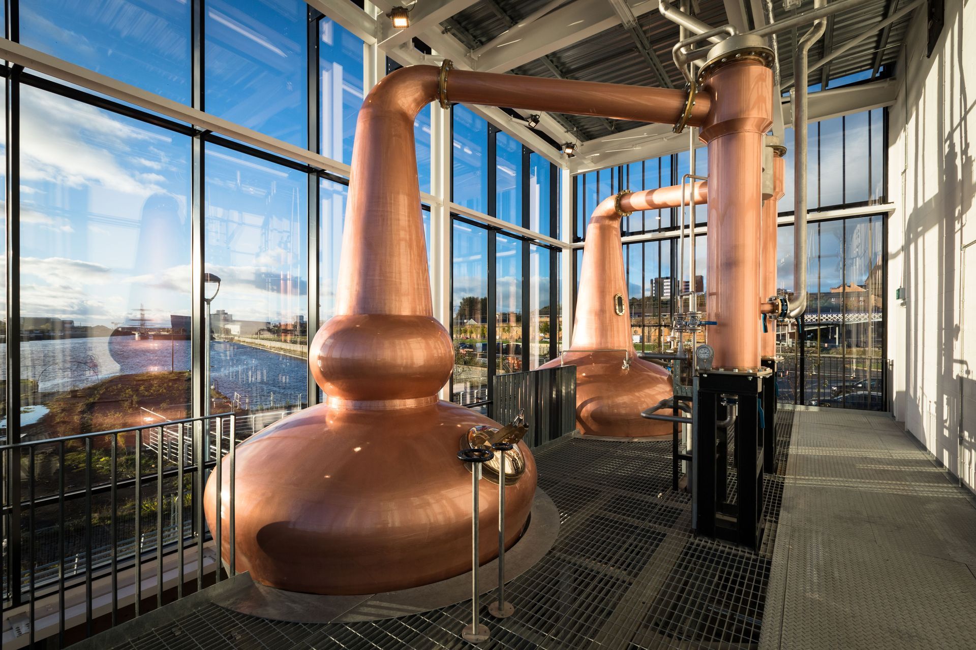 Copper stills in a distillery with a cityscape visible through windows.