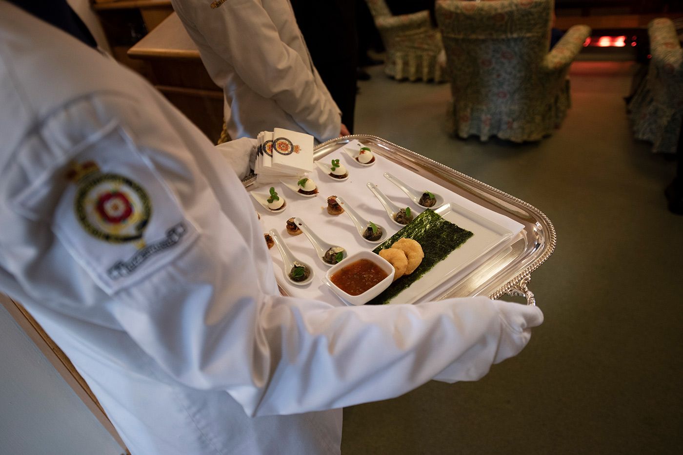 Server carrying a silver tray of appetizers: small bites on spoons, sauce, and seaweed.