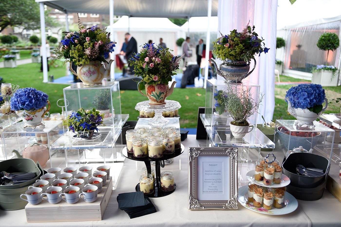 Dessert buffet with floral arrangements, cupcakes, and tea cups on a white table outdoors with tents in the background.