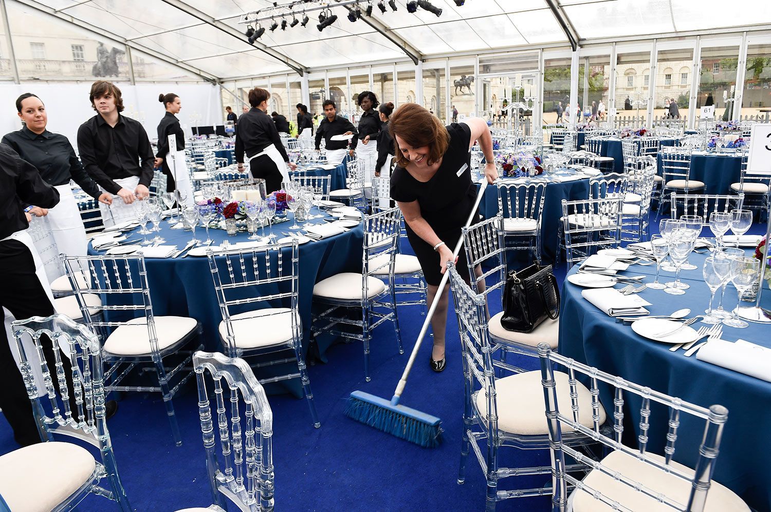 A woman sweeps a blue carpet in a large tent, surrounded by tables set for a formal event.