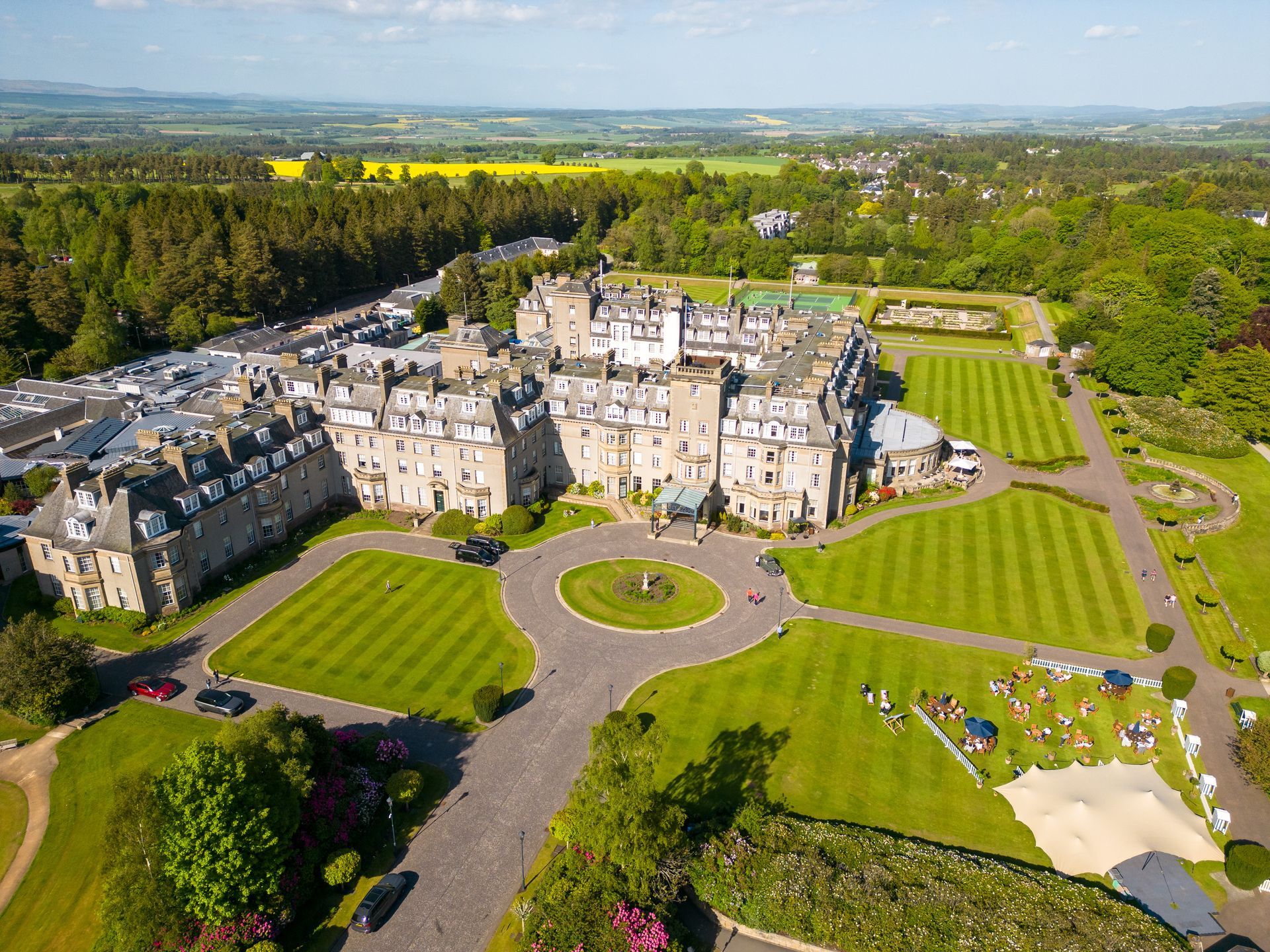 Aerial view of a large, stone hotel with green lawns and a roundabout entrance in a wooded area.