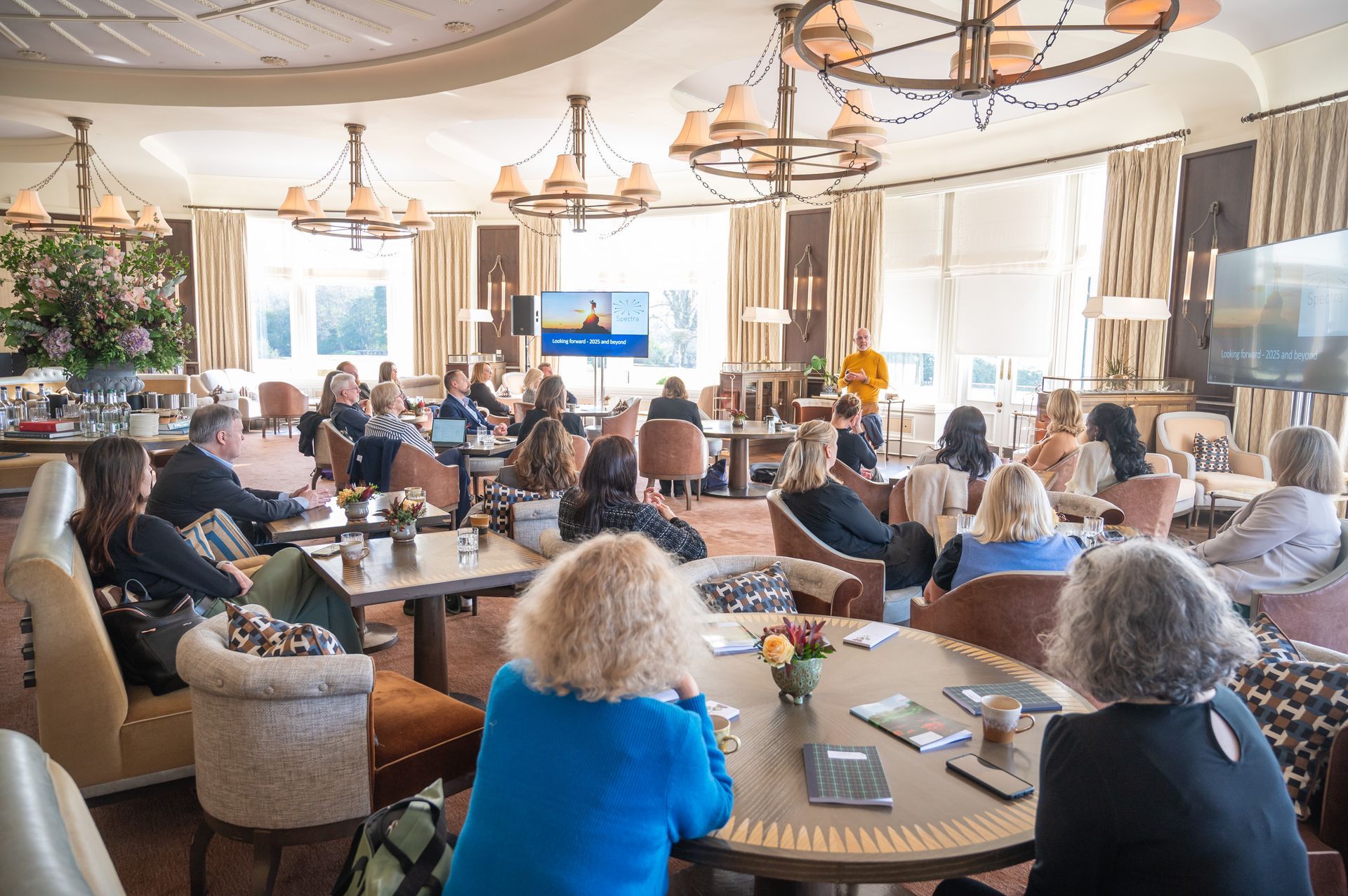 People seated at tables in a bright room, listening to a presentation on screens.
