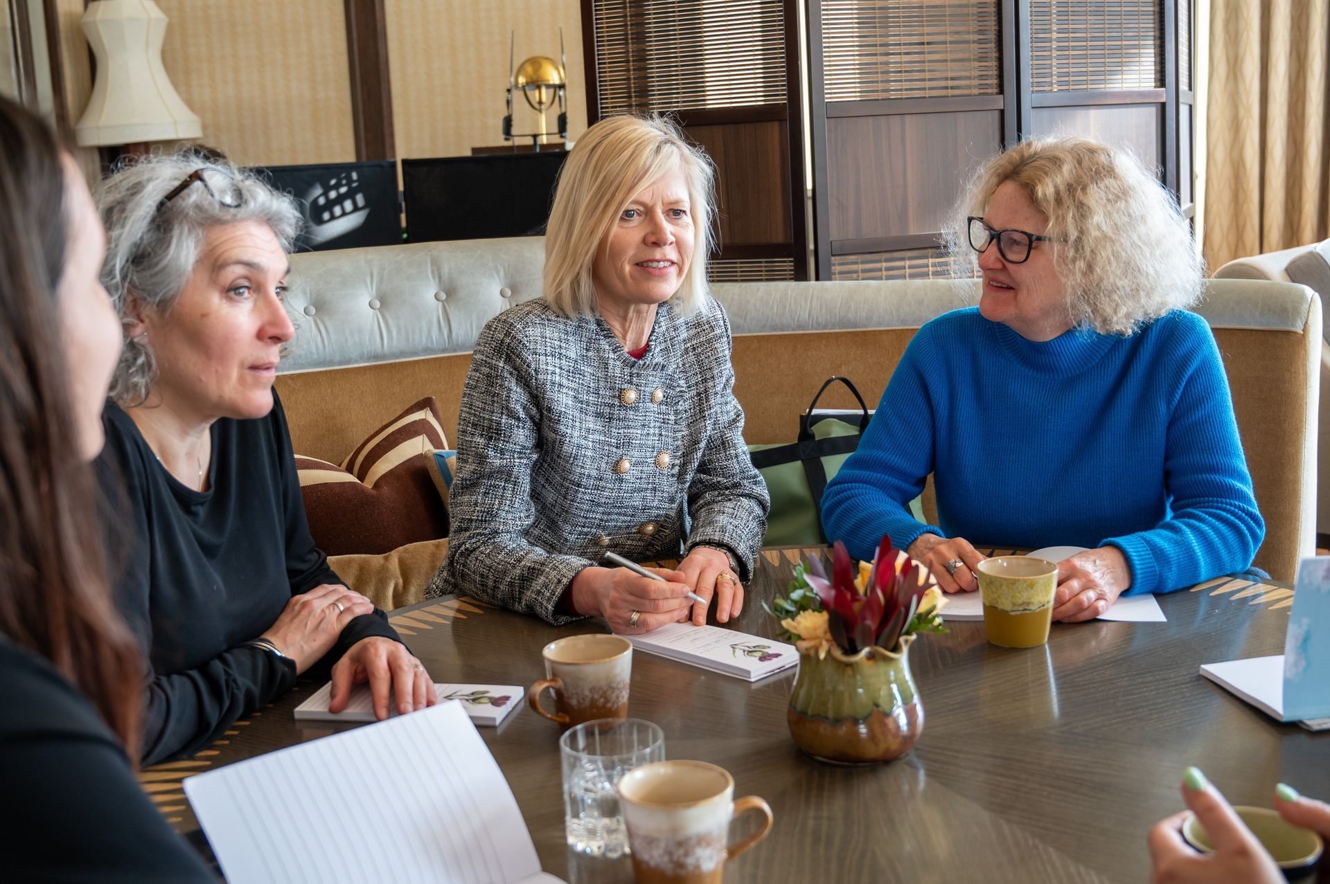 Four women seated around a table, engaged in conversation. Light-filled room, notebooks, cups, and flowers.