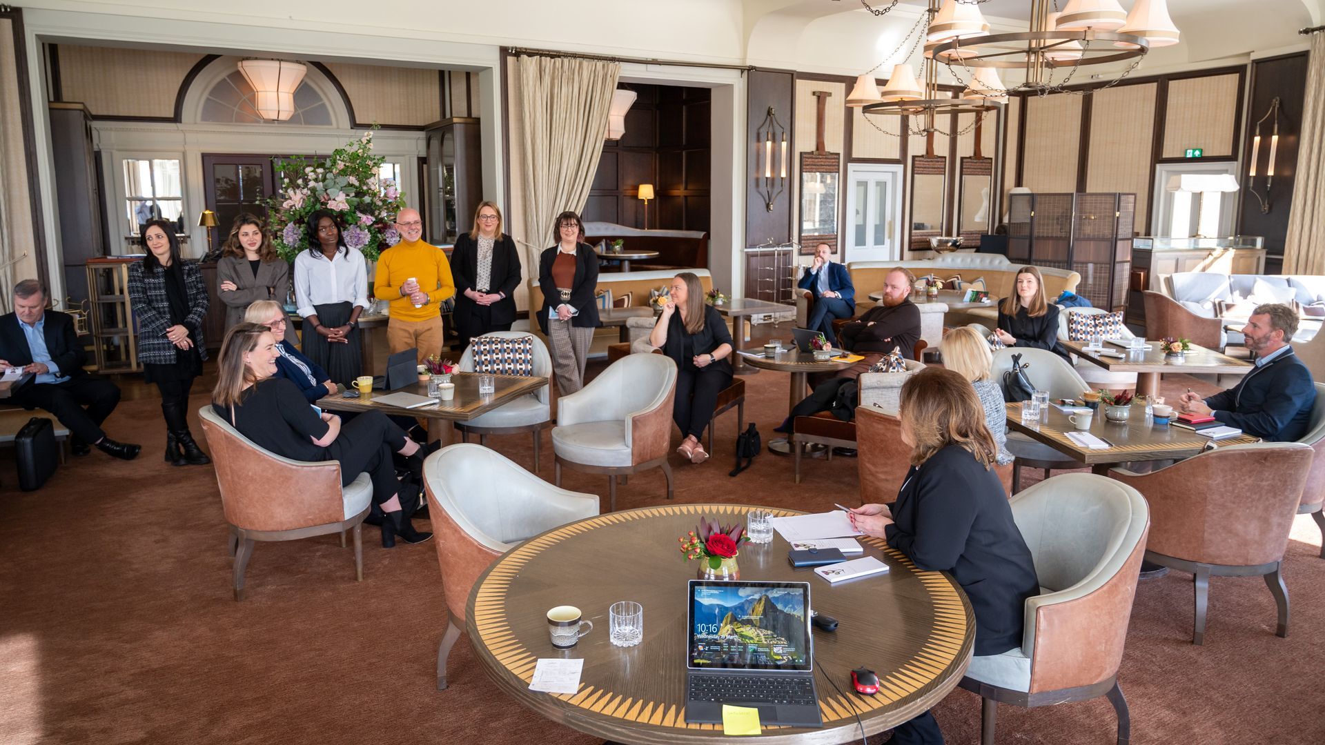 A group of people in a hotel meeting room. Some seated at tables, others standing. Laptop on table.