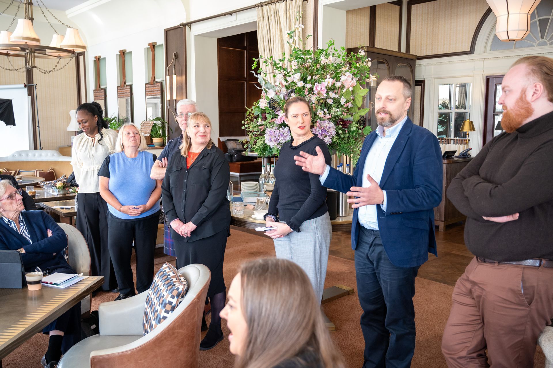 Group of people in a restaurant setting. A man in a blue blazer gestures as he speaks to the group.