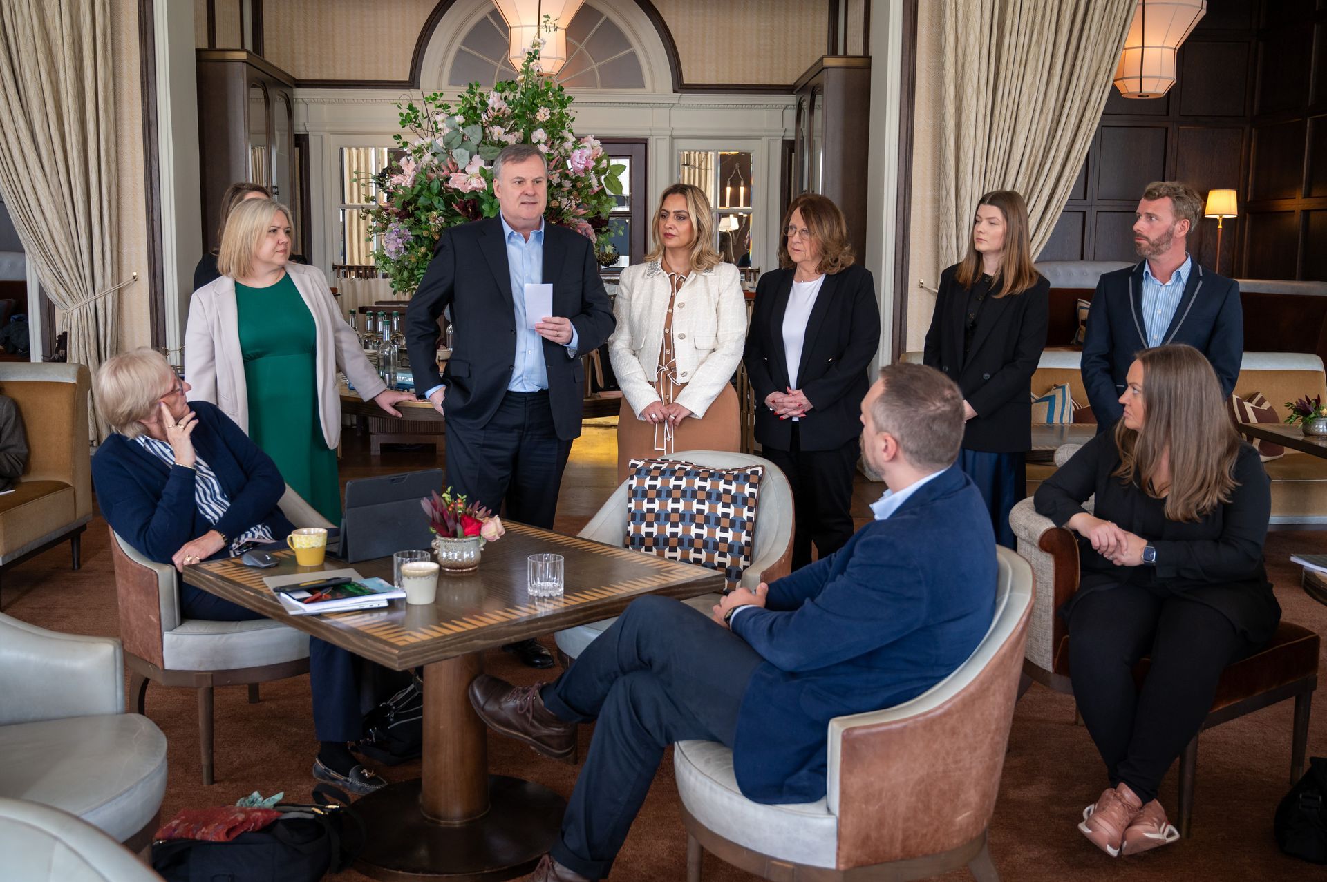 A group of people are gathered around a table in a hotel lobby, some listening, some speaking.
