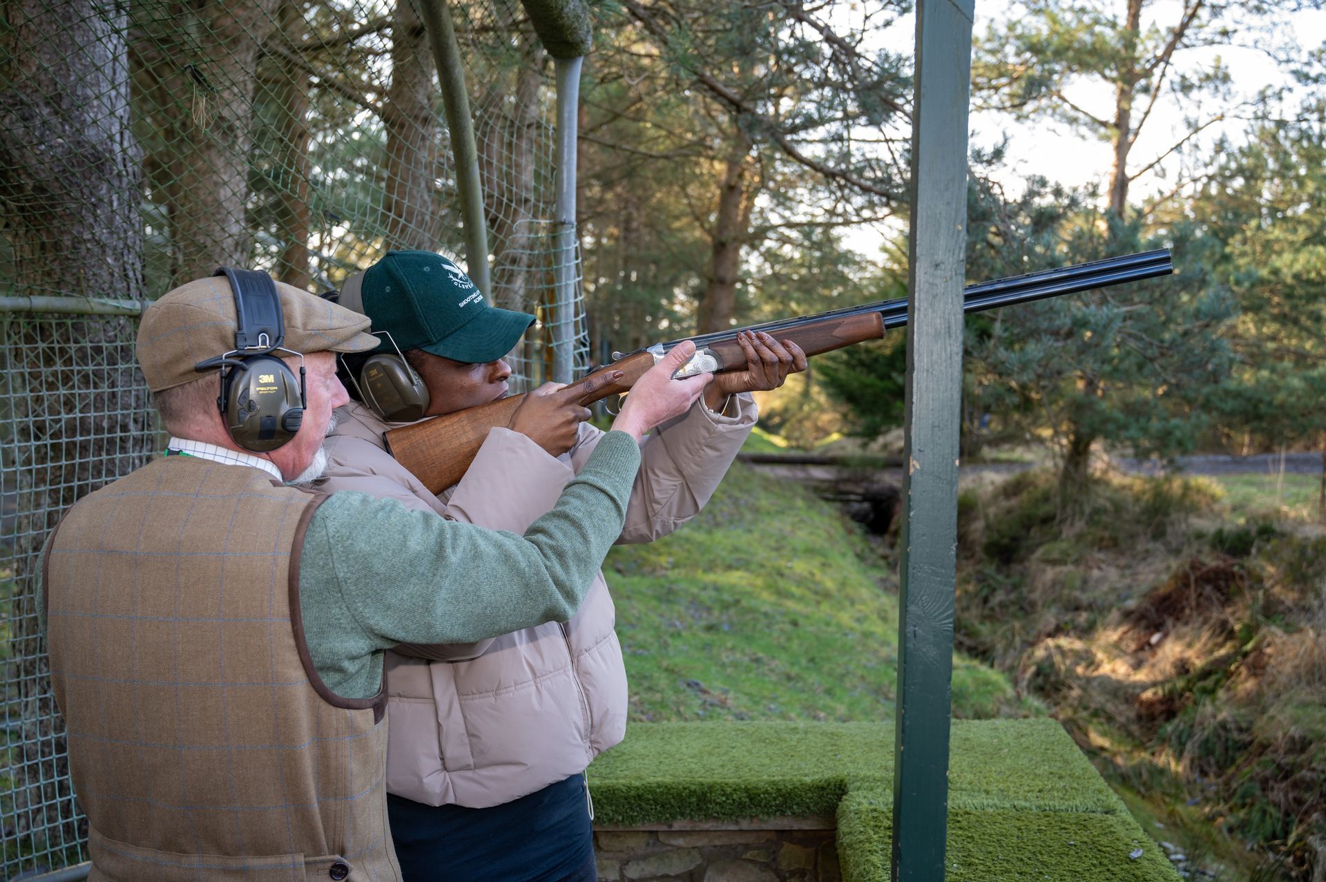 Man instructing person holding a shotgun outdoors, aiming at a target. Both wear hearing protection.